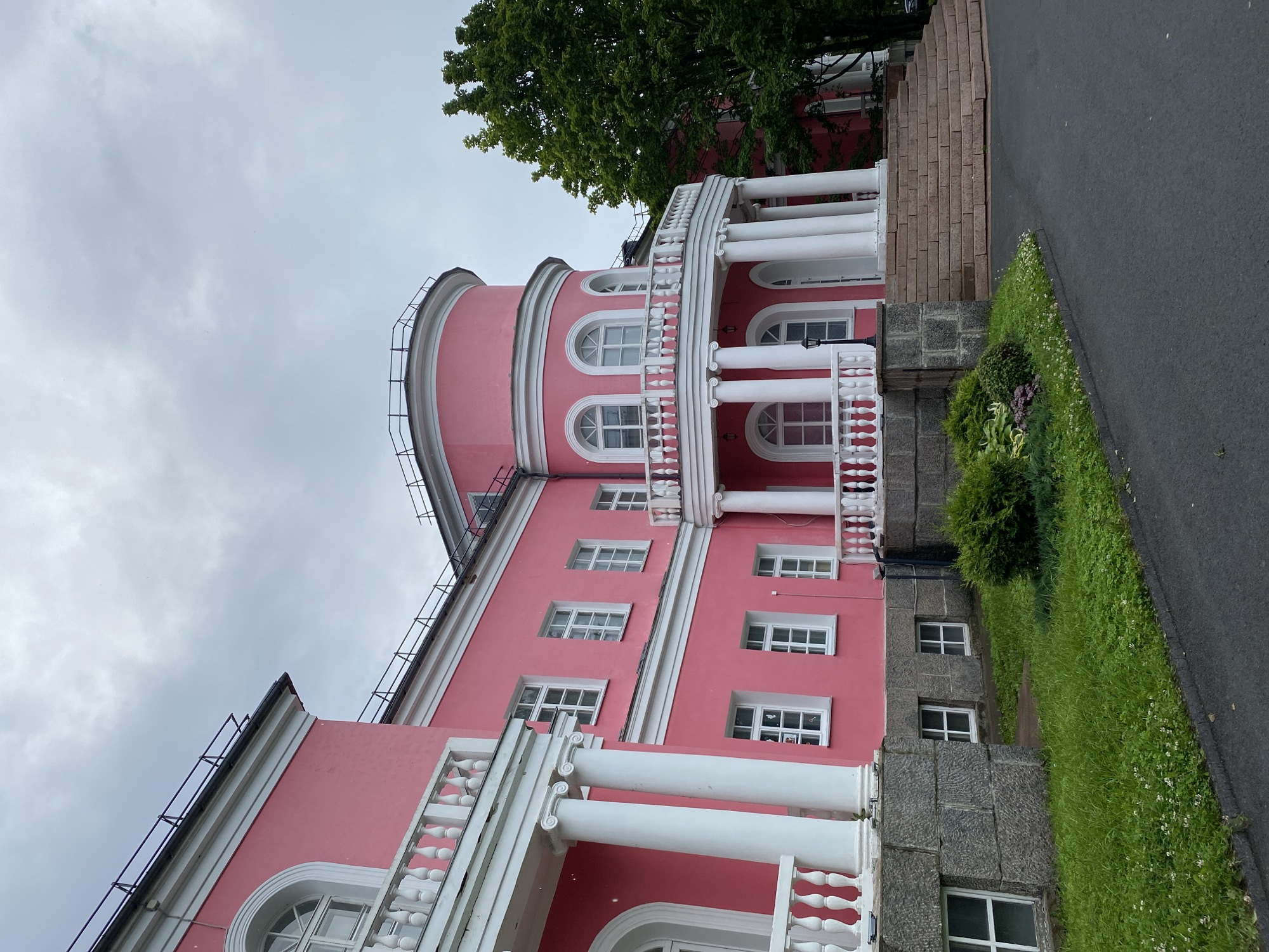 The image shows a historic pink building with white columns and railings in Petrozavodsk, Russia. The building has a semicircular section with arched windows and a balcony supported by columns. There are steps leading up to the entrance, surrounded by a manicured lawn and bushes. A large green tree stands to the right of the building. The sky is overcast with gray clouds. The scene is devoid of any people or discernible activity. The overall impression is one of a grand, albeit somewhat weathered, architectural landmark on a cloudy day.