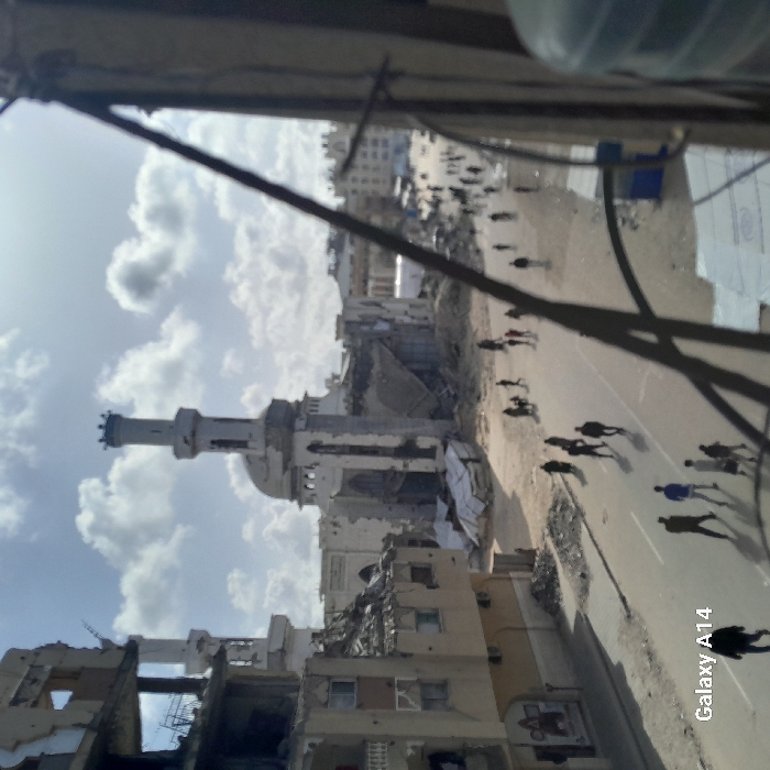The image shows a scene of devastation, likely a war zone. A partially destroyed mosque with a prominent minaret stands amidst damaged buildings. Debris litters the streets, and people are seen walking, suggesting some level of activity despite the destruction. The photo is taken from an elevated perspective, possibly from within another building, adding to the sense of observation of the aftermath. The sky is visible with white clouds.