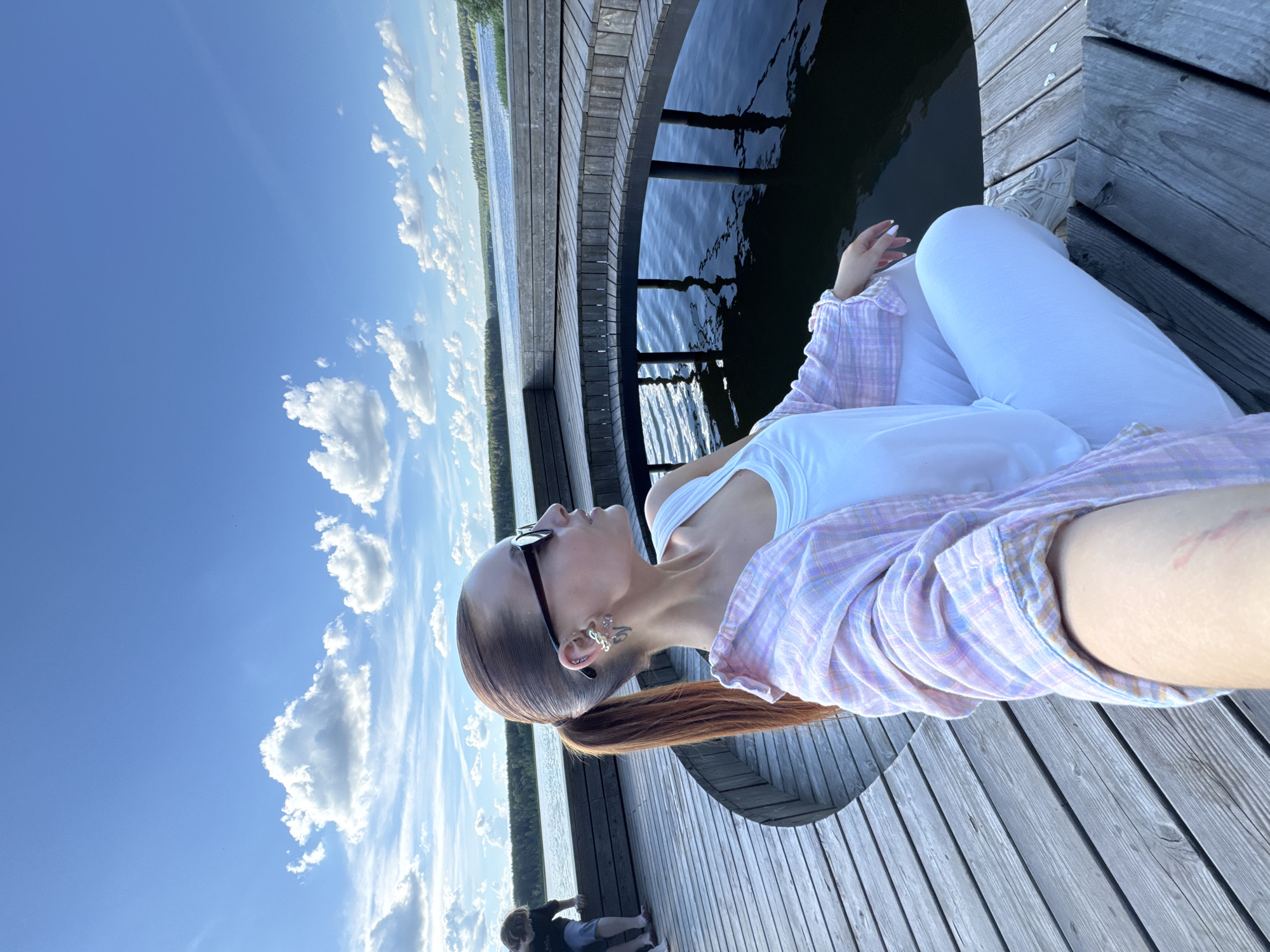 An overhead, slightly forward-facing, full shot captures a young woman with her hair in a ponytail, wearing sunglasses, a white tank top, a pink and white plaid shirt, and white pants. She is sitting on a wooden deck, which curves around a circular pool of dark water. The deck is made of weathered planks. In the background, a body of water stretches towards a treed horizon under a bright blue sky dotted with fluffy white clouds. To the left, a person in a dark t-shirt and shorts is partially visible, leaning on the deck. The woman is looking away from the camera towards the water. The scene appears to be set on a pier or waterfront area.