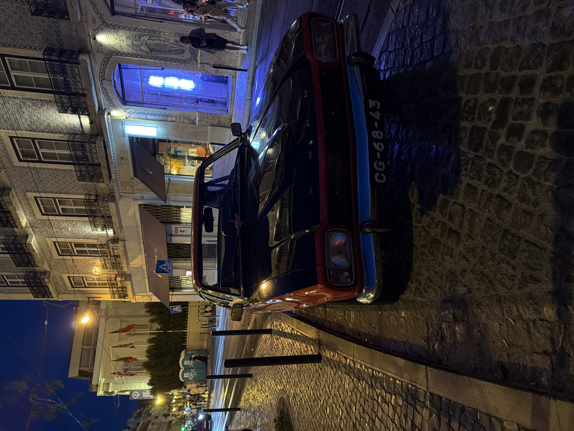 The image shows a vintage Ford Capri parked on a cobblestone street at night, likely in Lisbon, Portugal. The car is dark-colored with red trim and a blue stripe on the front bumper, bearing the license plate CG-68-43. In the background, a building with ornate balconies and tiled facade is visible, with storefronts featuring neon lights. Several people are walking on the sidewalk, and some portable toilets are seen on the left side of the street. The street is illuminated by streetlights and the lights from the buildings, casting shadows. The overall scene depicts a typical urban nighttime setting with a focus on the classic car.