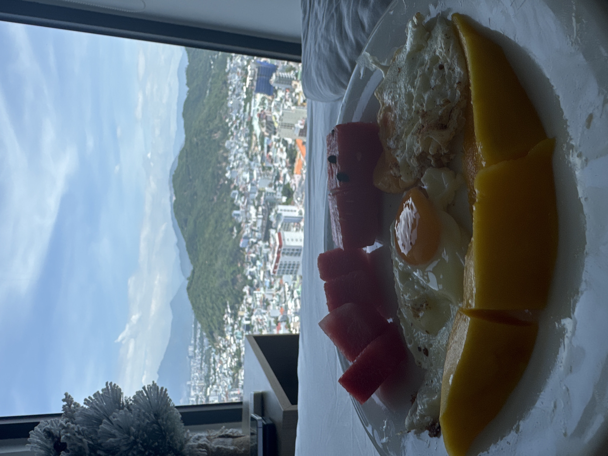 A close-up shot shows a plate of food in the foreground, with a city and mountain view in the background. The plate contains sliced watermelon, two fried eggs, and a large slice of mango. The food is placed on a white plate, which sits on a white surface, likely a bed. To the left, a decorative white, frosted object, resembling a small Christmas tree, is partially visible.
The background reveals a panoramic view of a city with numerous buildings, stretching towards rolling green hills and distant mountains. The sky is partly cloudy, with blue sky visible. The scene suggests a hotel room or apartment with a view, possibly located in Bac Nha Trang Ward, Vietnam, given the visual characteristics of the cityscape and terrain. No people are visible in the image.
