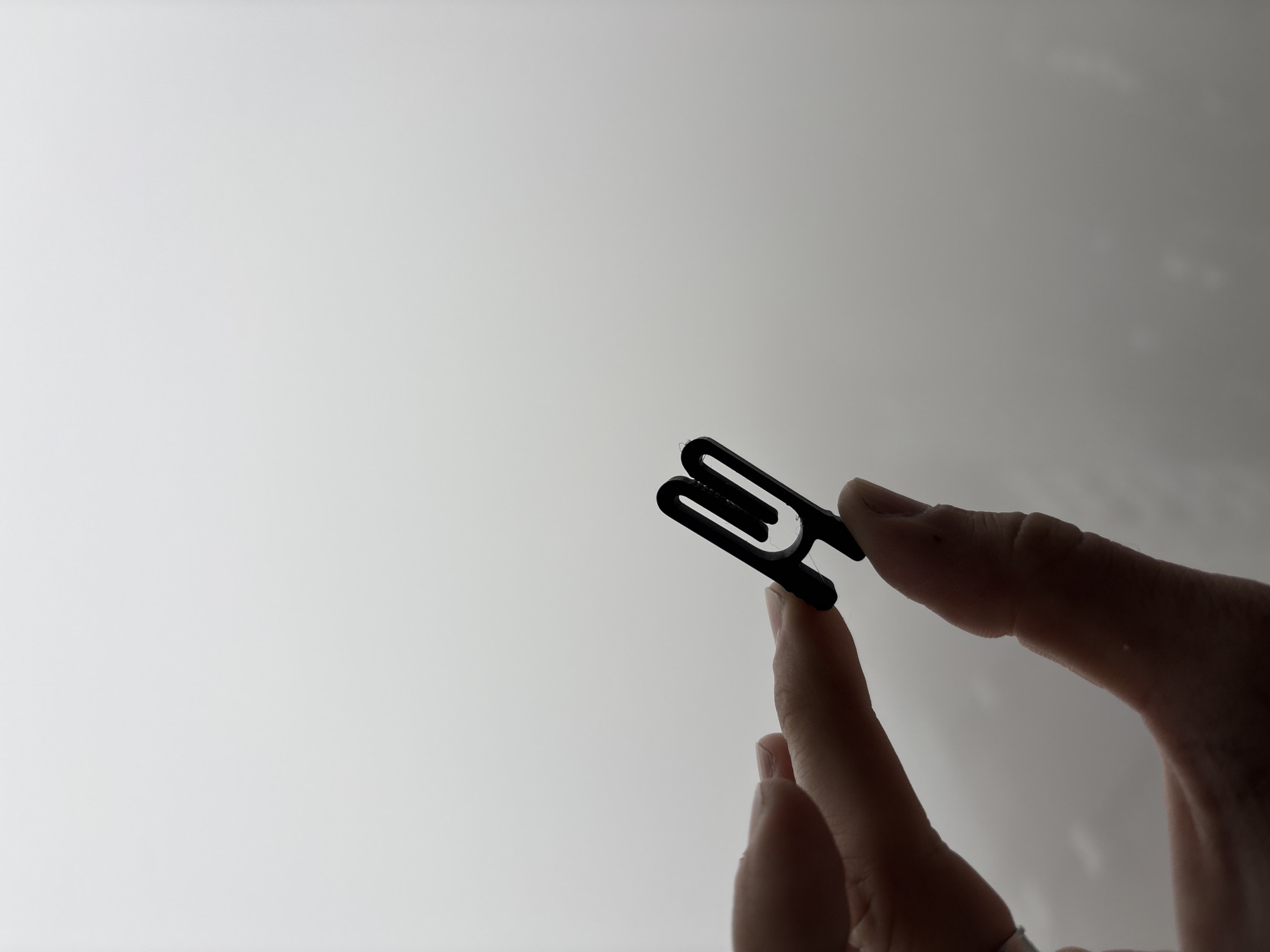 A person's fingers hold a black plastic clip against a hazy, light gray background. The clip is shaped like an 'H' with rounded ends. The fingers are silhouetted and appear to be light-skinned. The background is featureless and uniformly light gray, suggesting an overcast sky or a blurred indoor environment. There is no discernible urban landscape or specific location identifiable within the image.