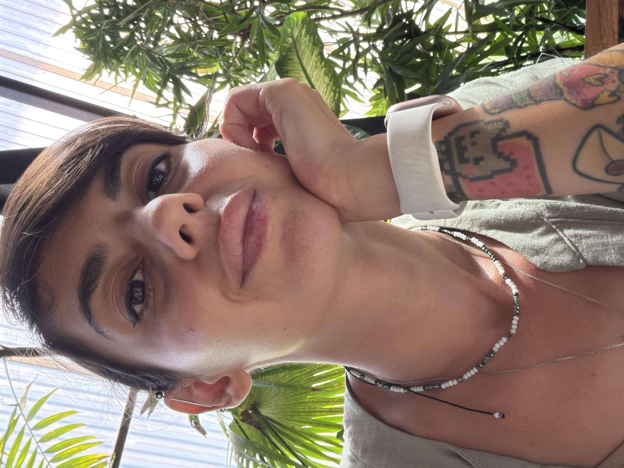 A woman of Mediterranean or Middle Eastern descent, with dark hair styled in a short bob and side-swept bangs, and prominent eyebrows, is captured in a close-up, selfie-style portrait. Her right hand rests on her cheek, supporting her chin. She is wearing a simple, light-colored V-neck top and a beaded necklace with white and green beads. A silver necklace is also visible underneath. On her left wrist, she wears a white smartwatch. Her left arm, visible in the frame, features colorful tattoos, including one that appears to be pixelated and another with a bottle-like design.
The background suggests an outdoor or semi-outdoor setting with lush green foliage from tropical plants. Sunlight streams through, creating bright highlights and casting dappled shadows. The environment appears to be in North Kuta, Indonesia, likely a restaurant or cafe with a garden setting, indicated by the visible overhead structure of what appears to be a transparent or translucent roof, and abundant plant life. The overall impression is a relaxed, casual moment, perhaps during daytime leisure.