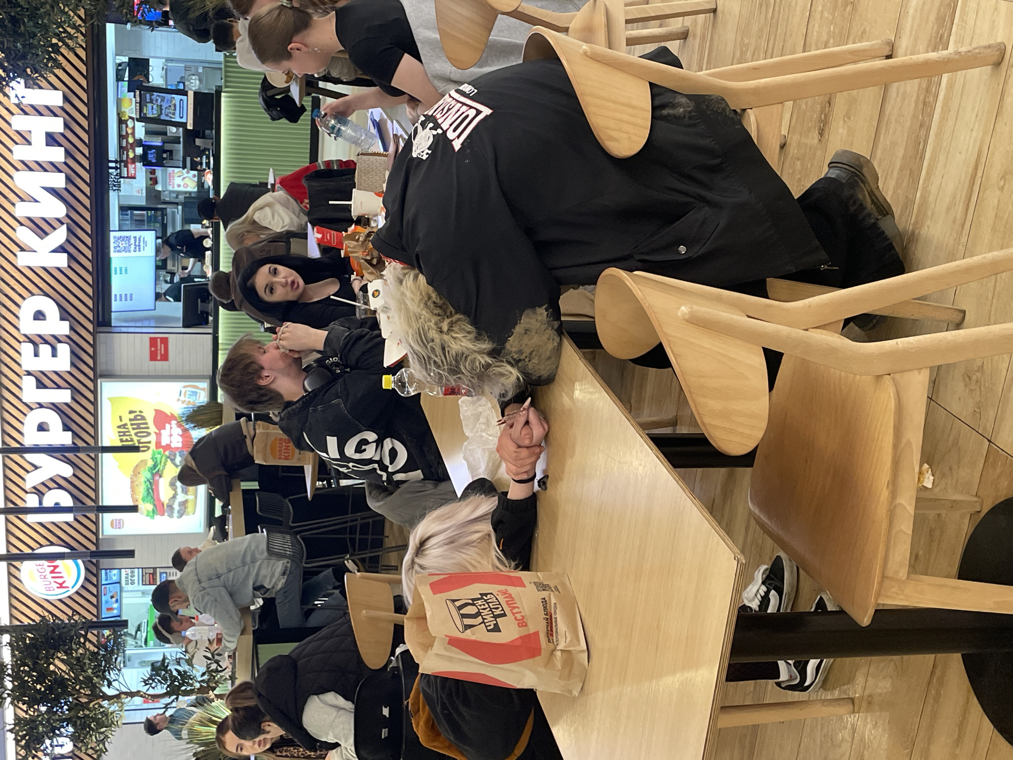 The image shows a group of people sitting at tables in a Burger King restaurant in Moscow, Russia. The restaurant's signage is visible, along with menu boards and a promotional poster. Several people are eating and talking, while others are on their phones. A woman with black hair is looking up in the direction of the camera. The tables are light wood. The floor is made of wide planks and also appears to be light wood.