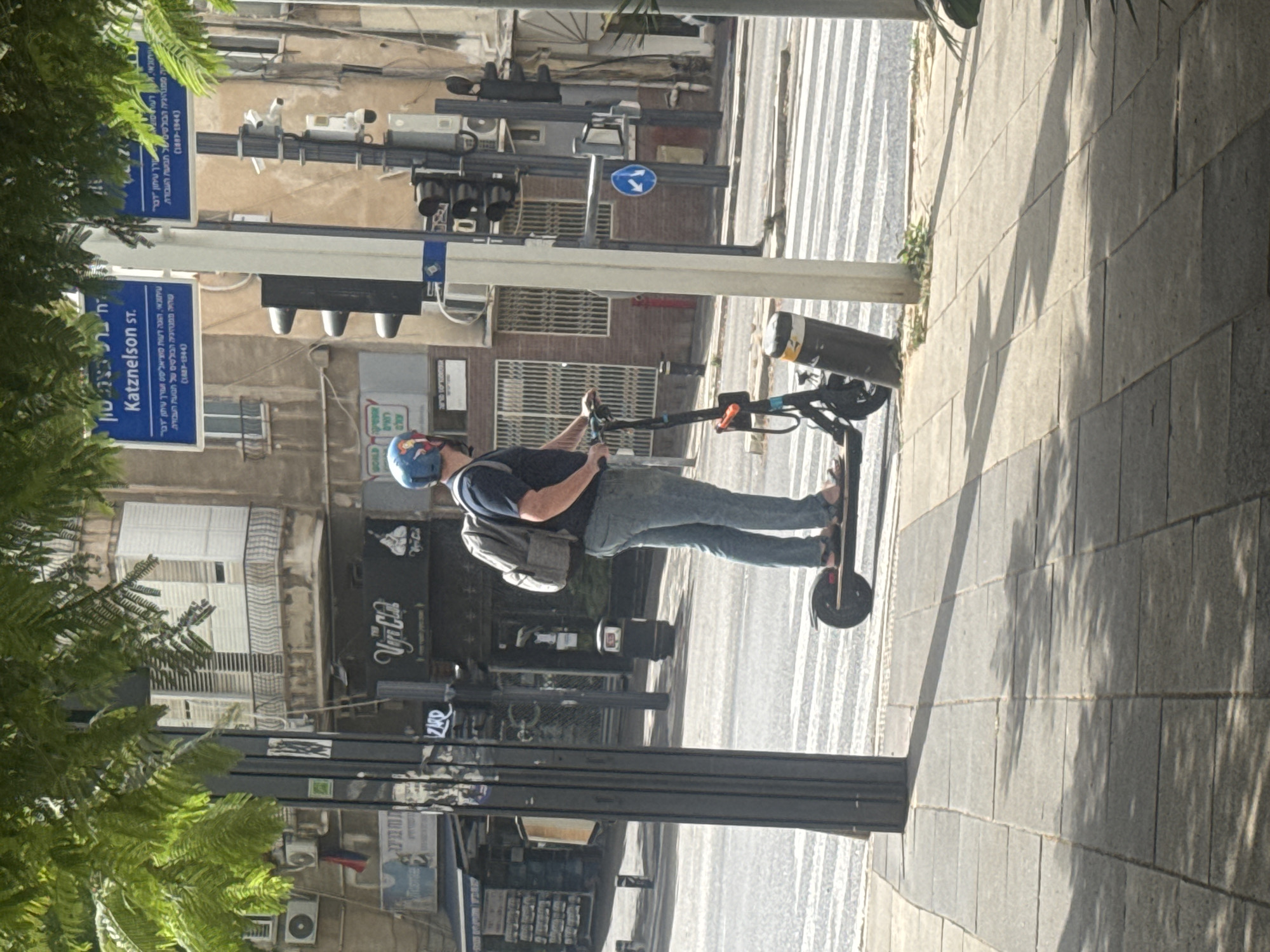 The image shows a man riding an electric scooter on a city street. He is wearing a blue helmet and a gray backpack. The street is lined with buildings and traffic lights. A street sign identifies the location as Katznelson Street in Givatayim, Israel. The sign also refers to a notable newspaper called "Davar" and a person called "Katznelson" (1887-1944). A restaurant is also visible behind the man on the scooter. The sidewalk is made of gray paving stones, with trees partially obstructing the left side of the image.