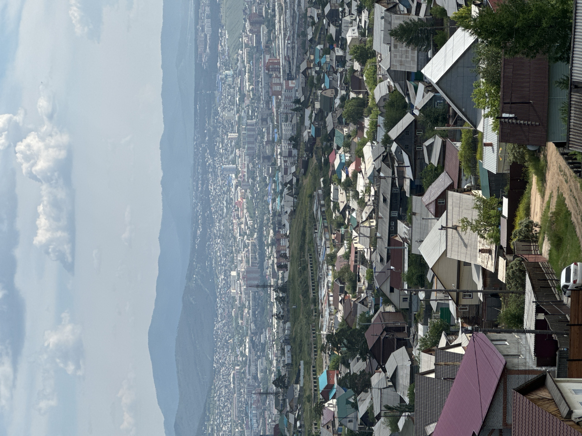 The image depicts an aerial view of a densely populated residential area in Ulan-Ude, Russia, likely a suburb or a hillside district, with a city skyline and mountains in the background. The foreground is dominated by numerous houses, characterized by their varied rooflines and colors, many of which are made of corrugated metal. The architecture suggests a mix of detached houses with small yards and what appear to be garages or outbuildings. A dirt road with a white car parked on it is visible in the lower right portion of the frame. In the middle ground, a larger urban area with taller buildings, including apartment blocks and some industrial structures, stretches across the landscape. Transmission towers are also present. The background is defined by rolling hills and mountains, partially obscured by a hazy atmosphere. The sky is partly cloudy, with cumulus clouds scattered across a light blue expanse. The overall impression is one of a sprawling settlement built on uneven terrain, merging into a more developed urban center.