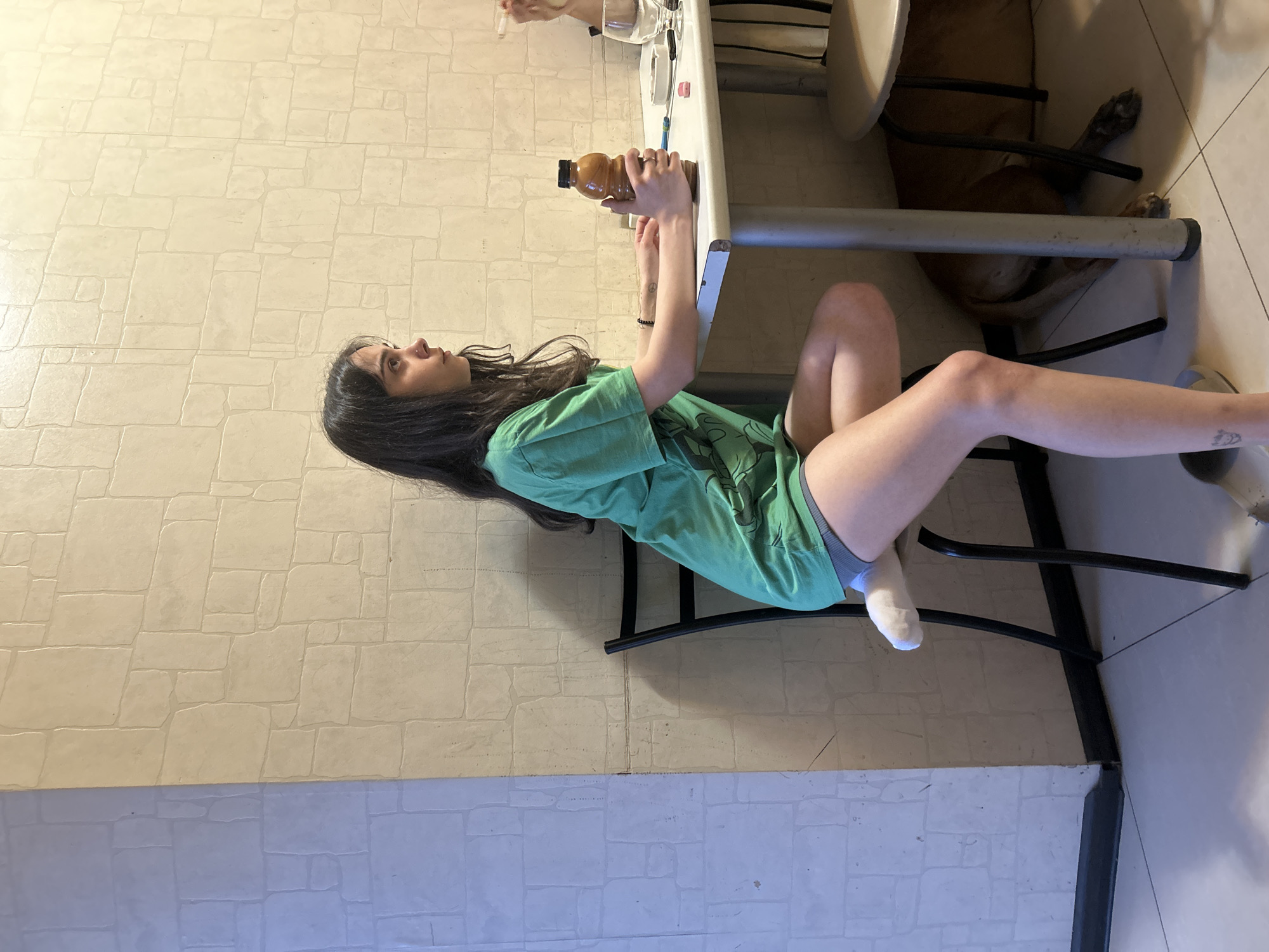 An overhead indoor shot shows a young woman sitting at a table, holding a bottle of brown liquid. She is wearing a green t-shirt and grey shorts, with one white sock visible on her left foot. The table is grey and appears to be made of metal. There are other items on the table, including a glass, an ashtray, a cigarette, and a pink eraser. To the right of the woman, another person is visible, holding a cigarette. A brown dog is lying on the floor underneath the table. The background consists of a beige wall with a stone-like pattern. The flooring is tiled.