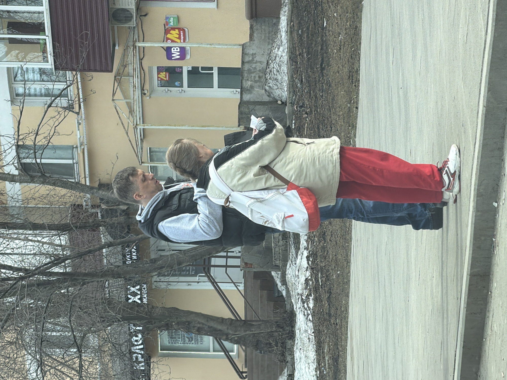 Two individuals stand close on a sidewalk in what appears to be Perm, Russia. A man in a grey hoodie and dark vest faces a woman, who is slightly shorter, in a light jacket and red pants. The woman seems to be holding a piece of paper or a small book. They stand next to a street with bare trees on one side and a building with various signs in Russian on the other. A sidewalk runs alongside the street. The buildings visible suggest a commercial area with signs for stores and services.