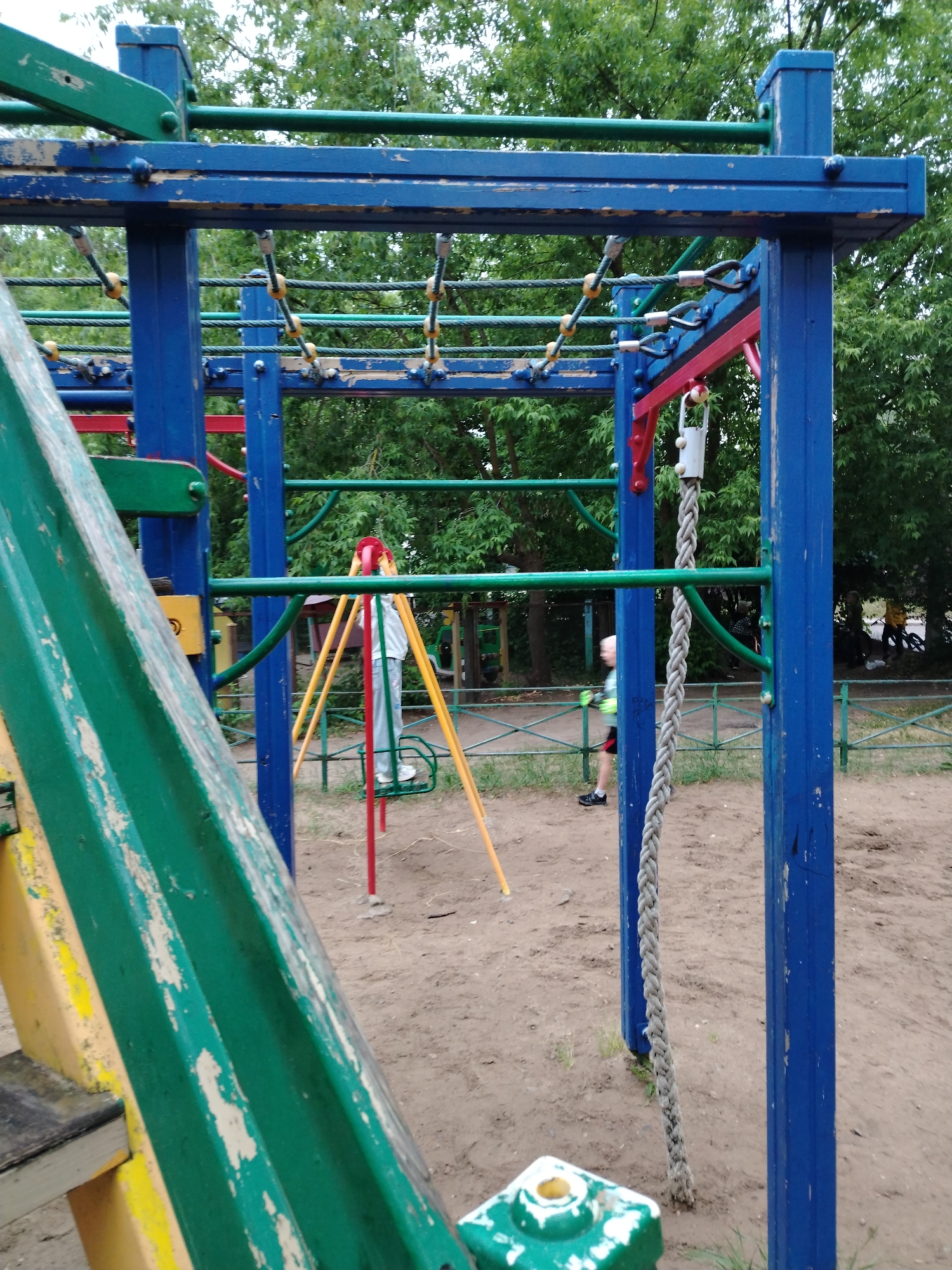 A playground with a blue metal structure, green slide, and rope is pictured on a cloudy day. The playground equipment is worn and shows signs of rust and peeling paint. A person in a white shirt and grey pants is standing near a yellow and red swing set in the background. Another person, blurred by motion, is running in front of the blue structure. The ground is covered in sand and dirt with some patches of grass. Trees are visible in the background, creating a natural setting.