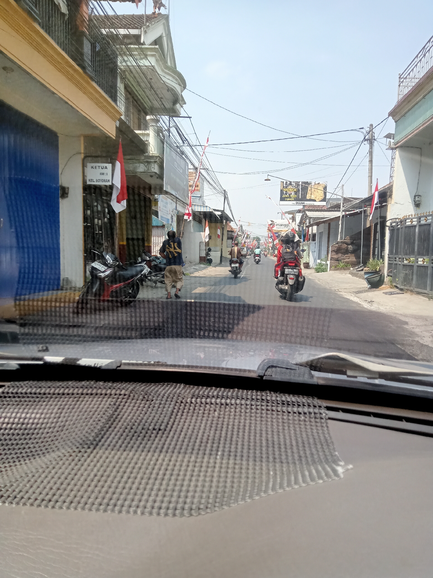 The image captures a street scene in Botoran, Indonesia, viewed from inside a vehicle. The street is lined with buildings, some with yellow and white facades, and others with light green accents. A blue garage door is visible on the left. Several motorcycles are parked along the side of the road, and a few are in motion on the street. Three individuals are on motorcycles: one wearing a red jacket and black helmet, another with a black shirt and backpack, and a third with a striped shirt. A person in a dark shirt and light-colored pants is walking away from the camera on the sidewalk. Indonesian flags are displayed on poles along the street. Overhead, a network of electrical wires crisscrosses the sky. The weather appears clear and sunny. The foreground shows the dashboard and windshield of the vehicle from which the photograph was taken.