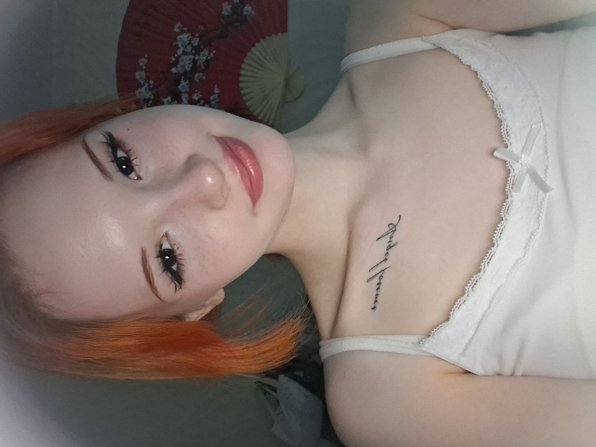 A close-up, slightly angled, indoor shot features a fair-skinned woman with red/orange hair, lying on her back, presumably in a private residence. Her facial features, including her eyes, nose, and lips, are clearly visible and made up. She wears a white camisole with lace trim, and a tattoo is visible on her chest. An opened, red and white floral fan is behind her. There is no indication of the location being in Chaikovsky, Russia.