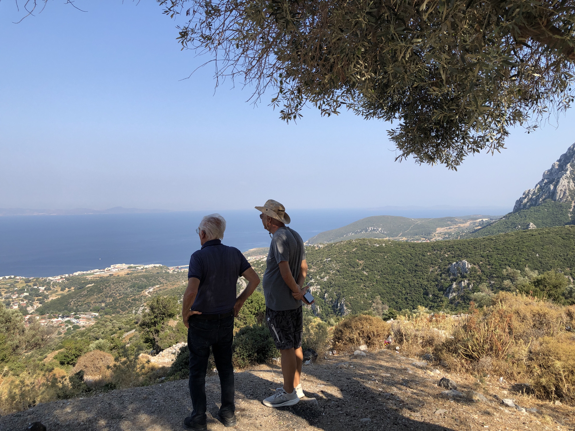 Two individuals are positioned on an elevated viewpoint overlooking a coastal landscape. The person on the left, facing away from the camera, wears a dark blue short-sleeved shirt and dark jeans. The person on the right, also facing away, wears a light gray short-sleeved shirt, patterned shorts, and a light-colored hat. They are standing side-by-side, looking out towards the horizon.
The environment consists of dry, scrubby vegetation in the foreground, with a mountainous, forested terrain stretching into the distance. A body of water, likely the sea, is visible beyond the populated coastal area. The sky is clear and blue. The scene appears to be captured during daylight hours, possibly in the late afternoon given the shadow patterns. The location is identified as being in the vicinity of Karaburun, Turkey.