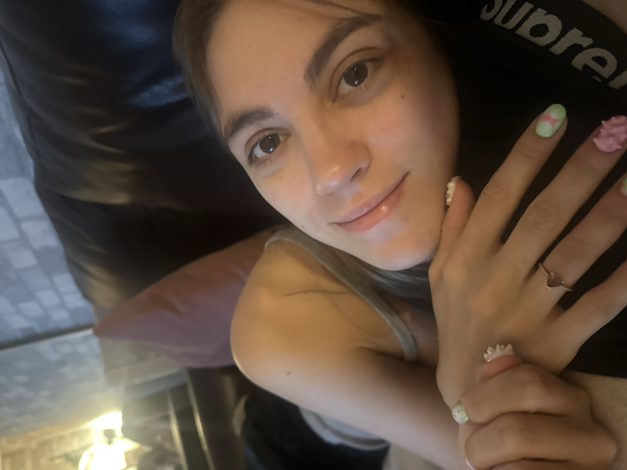 The image shows a close-up, slightly blurry, selfie of a young woman lying down. She has brown eyes, dark hair pulled back, and a light complexion. She is wearing a light-colored tank top. Her hands are in the foreground, resting on a black surface that appears to have the word "Supreme" printed on it. Her fingernails are decorated with colorful designs, including flowers and geometric patterns, and she is wearing a few rings. The background is dark and out of focus, but appears to be an indoor setting, possibly a couch with a pink pillow. There is no indication of the location being Moscow, Russia.