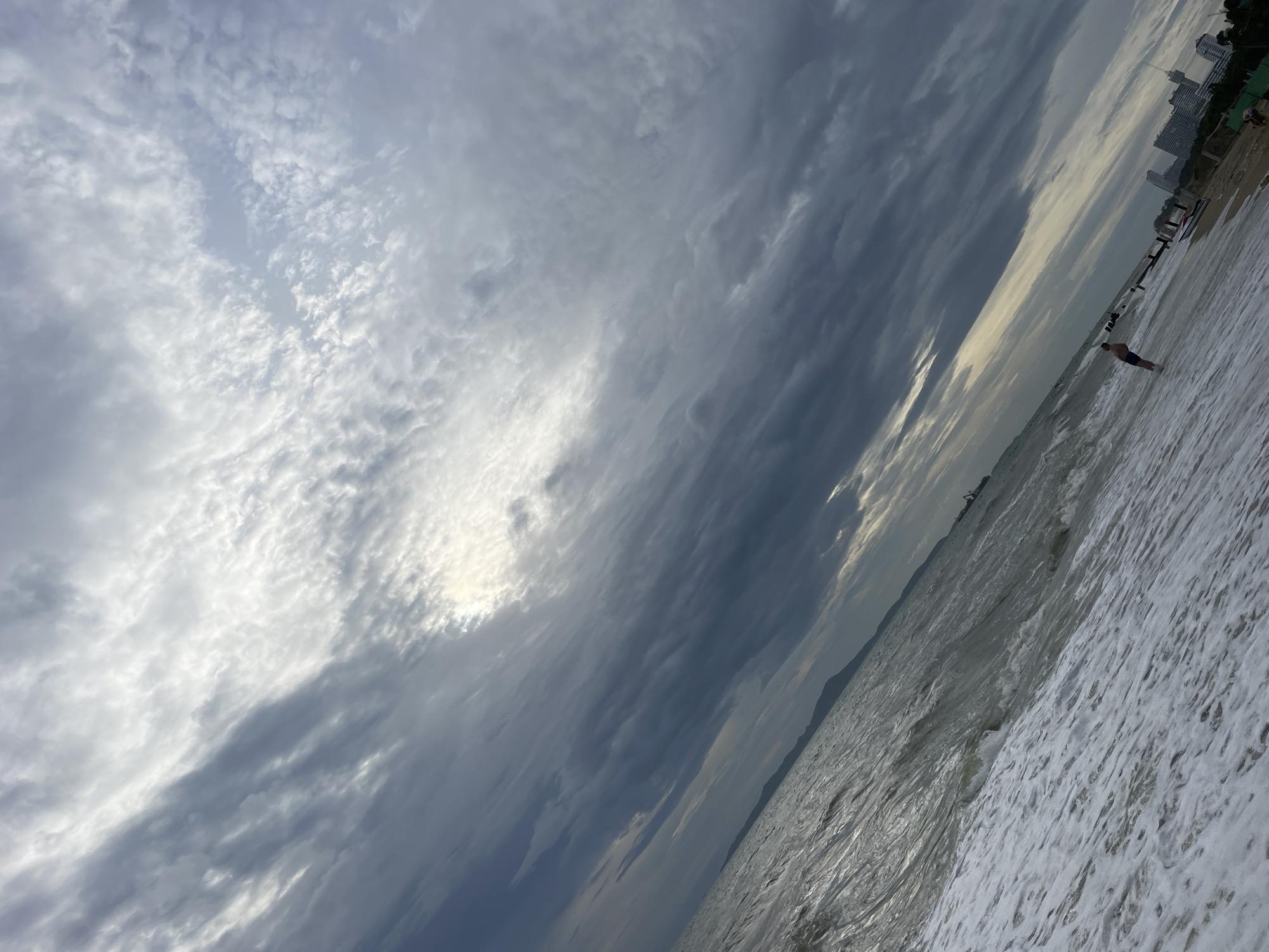 The image depicts a coastal scene in Pattaya City, Thailand, under an overcast sky with dramatic cloud formations. The foreground is dominated by foamy ocean waves washing onto a sandy beach. In the middle ground, a lone individual, appearing to be a man wearing dark swim trunks, stands partially submerged in the shallow water, facing away from the camera. Further in the distance, along the shoreline, several modern buildings, including what appear to be high-rise hotels or condominiums, are visible, as well as construction cranes, indicating ongoing development. The sea stretches towards the horizon, meeting the textured, cloudy sky. The image is captured at an angle, giving a sense of perspective and depth to the scene.