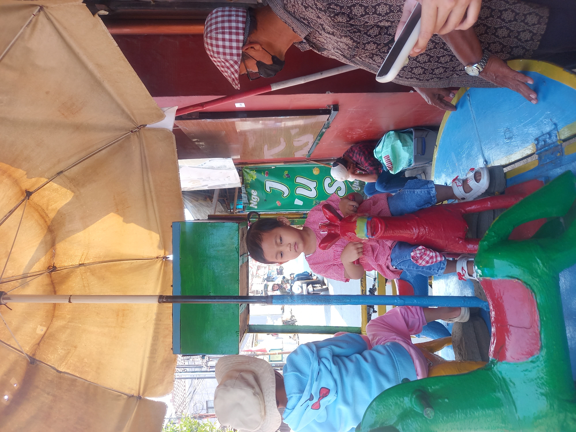 In Linggar, Indonesia, a carousel ride, possibly for children, is set up outdoors. A young girl in a red and white checkered shirt and blue jeans sits on a red rocking horse, her hands gripping its reins. To her left, another child wearing a blue hooded jacket and light-colored hat faces away from the camera, also seated on a colorful carousel animal. In the background, an adult male wearing a patterned shirt, a checkered cap, glasses, and a black face mask is partially visible, looking to the right. Another person in a red and black checkered shirt is seated further back, leaning against a red wall, with a light blue bag at their feet. The carousel is covered by a large, faded yellow umbrella providing shade. Signs indicating "JUS" are visible on a green board behind the children. The scene appears to be a public space with some urban elements visible in the background, such as buildings and a motorcycle.