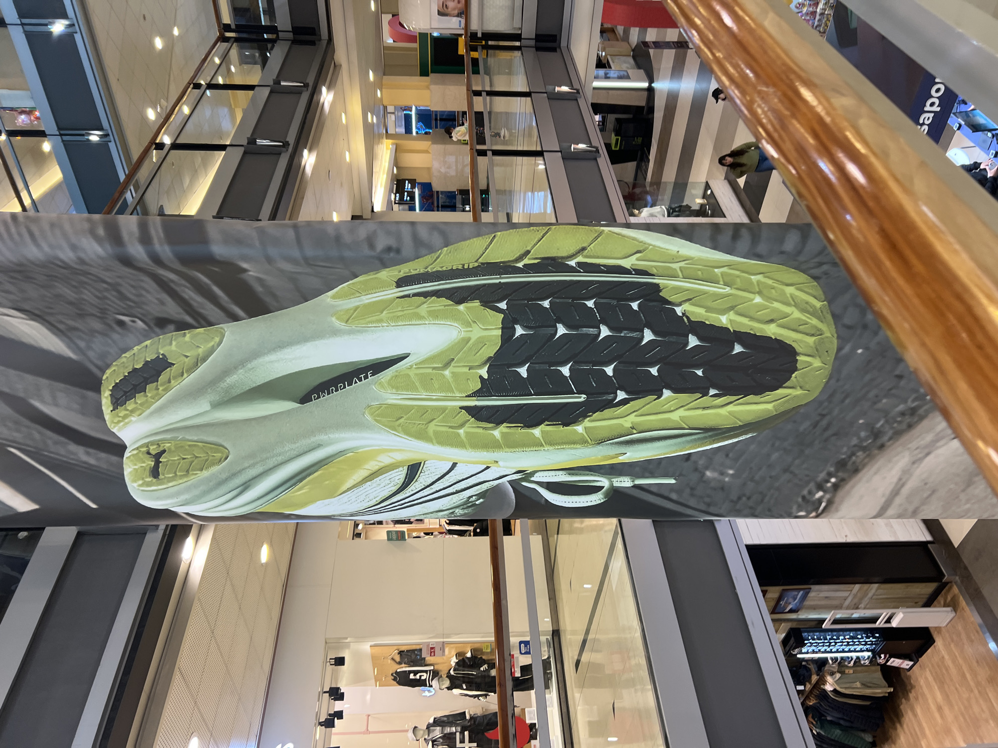 The image shows an advertisement of a running shoe in a shopping mall. The shoe is a light green and black color with a prominent sole. The advertisement is placed in a multi-level shopping mall, and escalators can be seen in the background. The location cannot be determined to be Buenos Aires.