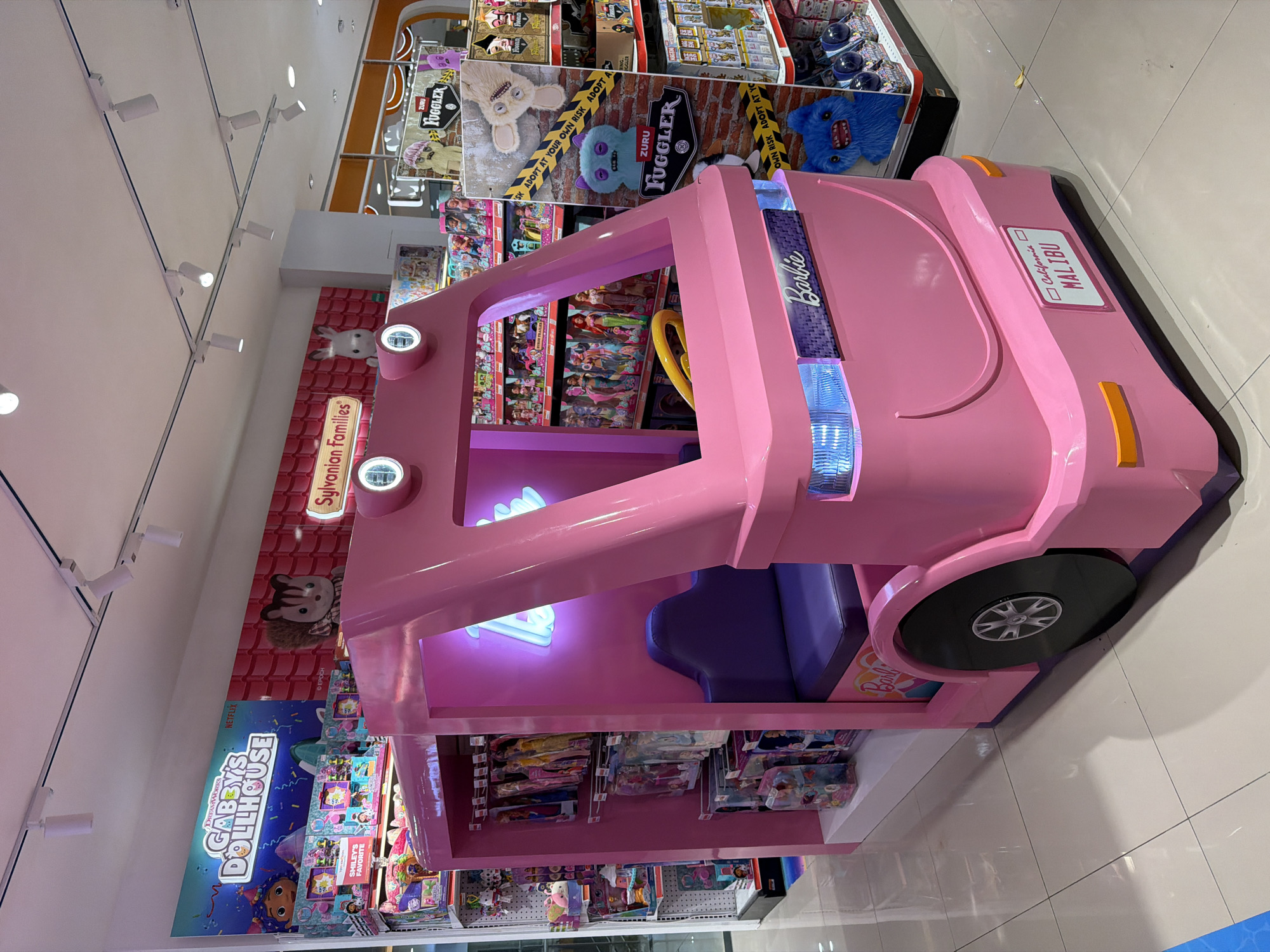 This indoor shot depicts a toy store, potentially in the city of Kuta, Indonesia. The focus is on a pink, life-size Barbie Malibu car display, complete with a seat and wheels. The car contains shelves filled with various Barbie dolls and related merchandise. Behind and around the Barbie display are shelves stocked with other popular toy brands such as "Sylvanian Families" and "Gabby's Dollhouse," as well as "Fuggler" toys from the brand ZURU. The environment suggests a well-lit retail setting, with emphasis on the toy section. No people are visible in the shot.