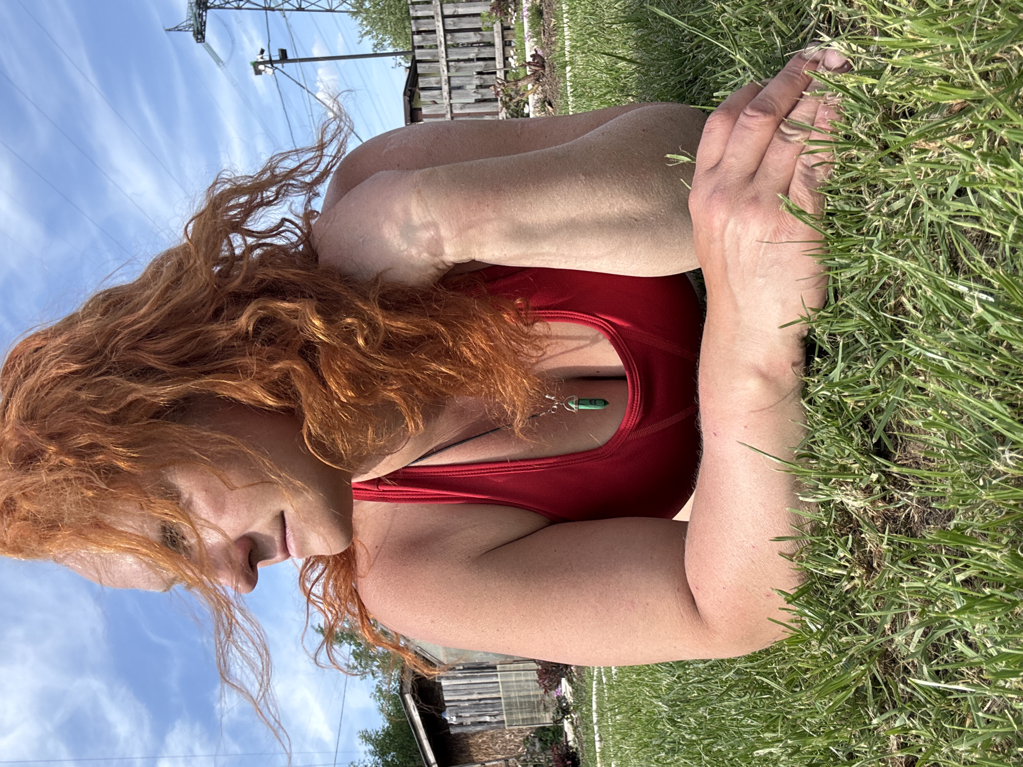 A woman with red hair and a red tank top is lying on a patch of grass. She is facing to the left. She has a necklace with a green stone. In the background is a wooden fence, a utility pole, a blue sky with clouds, and possibly a building. The location cannot be determined as Chaikovsky, Russia, as the photo does not contain any contextual information related to the city.