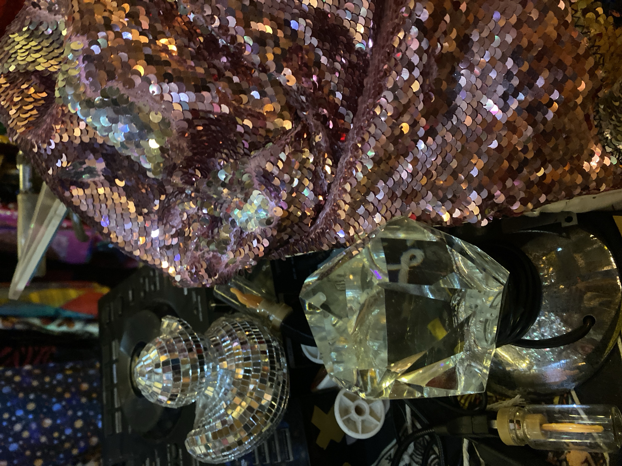 The image shows a close-up of various objects. A section of fabric covered in iridescent sequins is at the top of the frame. Below the sequins is a small mirrored disco ball, a clear geometric prism, and a silver lamp base. A small glass bottle with a filament bulb is also visible. The environment appears cluttered. The location is Baan Nai Suan, Thailand.