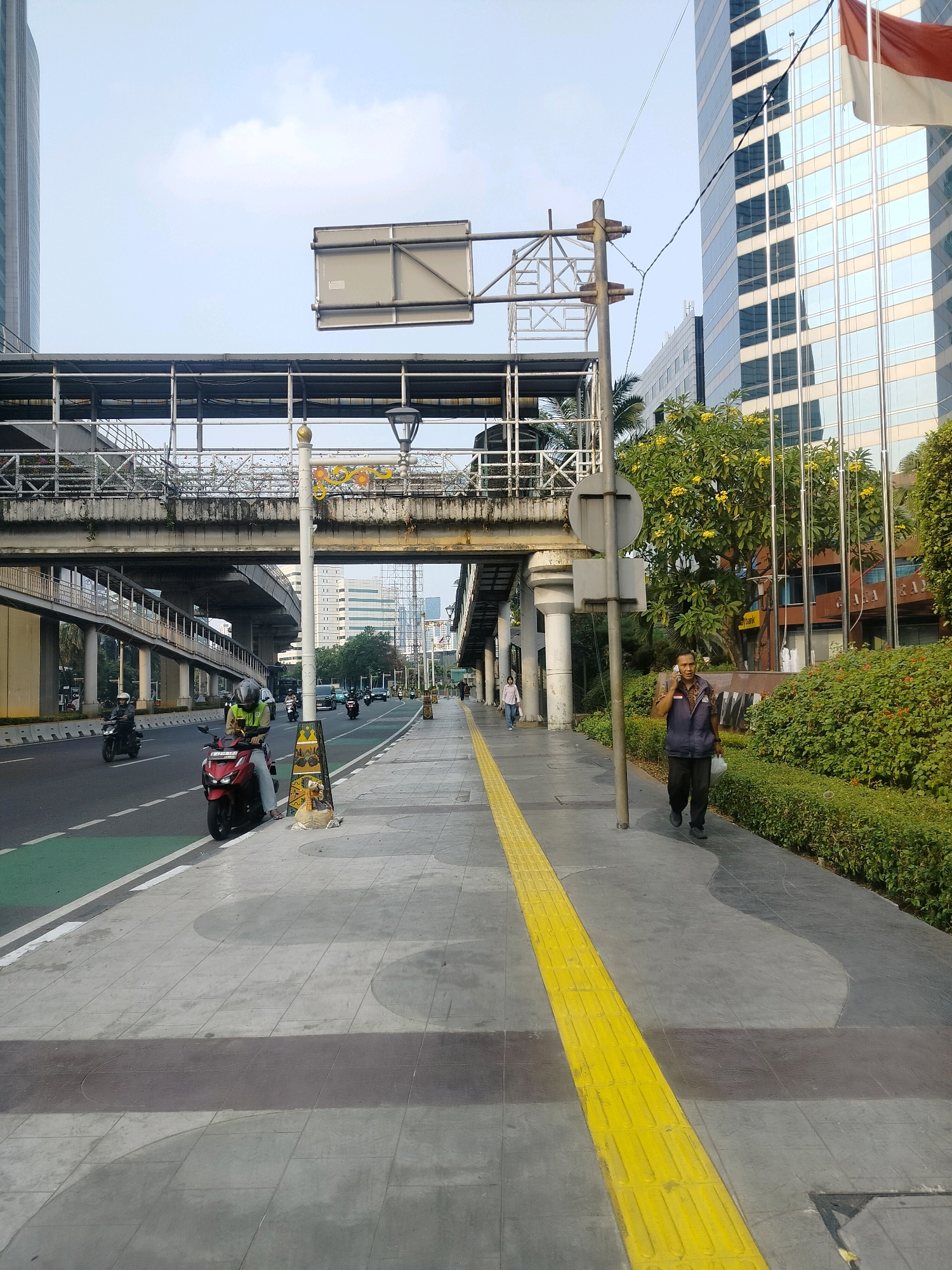 The image shows a street scene in South Jakarta, Indonesia, likely in the business district. A man in a dark blue vest and dark pants is walking on a sidewalk, talking on his phone and carrying a white bag. The sidewalk has a tactile paving strip, indicating it is accessible for visually impaired pedestrians. Behind the man, there is a dense green hedge and a modern glass-fronted building.
In the foreground, a red motorcycle is parked on the side of the road, with a person wearing a lime green vest and helmet sitting on it. The road has multiple lanes, with some vehicles, including cars and motorcycles, visible in the distance. A green bicycle lane is marked on the left side of the road.
An elevated pedestrian bridge or walkway stretches across the scene, supported by concrete pillars. Above the pedestrian bridge, there are signs and streetlights. The sky is bright and clear, suggesting it is daytime, possibly late afternoon given the lighting. The overall environment is urban, with elements of modern infrastructure, green spaces, and active transportation.