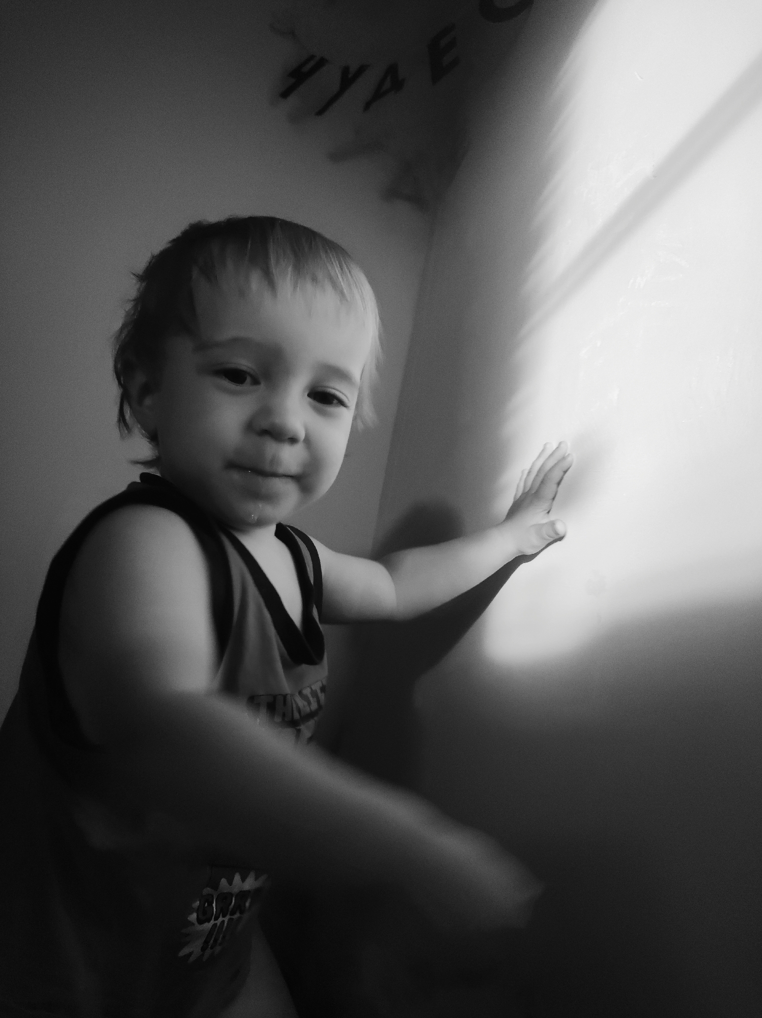 This is a black and white, low-light, close-up photograph of a young child. The child appears to be a toddler or very young child, with light-colored hair and dark eyes. They are wearing a sleeveless shirt with a dark trim. The child is positioned to the left of the frame, facing towards the right, and has their right hand extended to touch a brightly lit wall. The light source appears to be from the right, casting a strong shadow of the child's arm and hand onto the wall. The child has a curious or slightly mischievous expression, with a hint of liquid, possibly saliva, at the corner of their mouth.
In the upper left background, there is some faint text on the wall, which appears to be in Cyrillic letters. Some of the visible characters are "У", "Д", "Е", and possibly "Я" or "Ч". The overall mood of the image is intimate and captures a moment of childhood exploration and wonder. The setting seems to be indoors, likely a room in a home, given the wall and the close-up framing. The time of day is indeterminate due to the artificial lighting, but the shadows suggest a deliberate lighting setup rather than natural daylight. The location context is specified as Фоки, Russia.