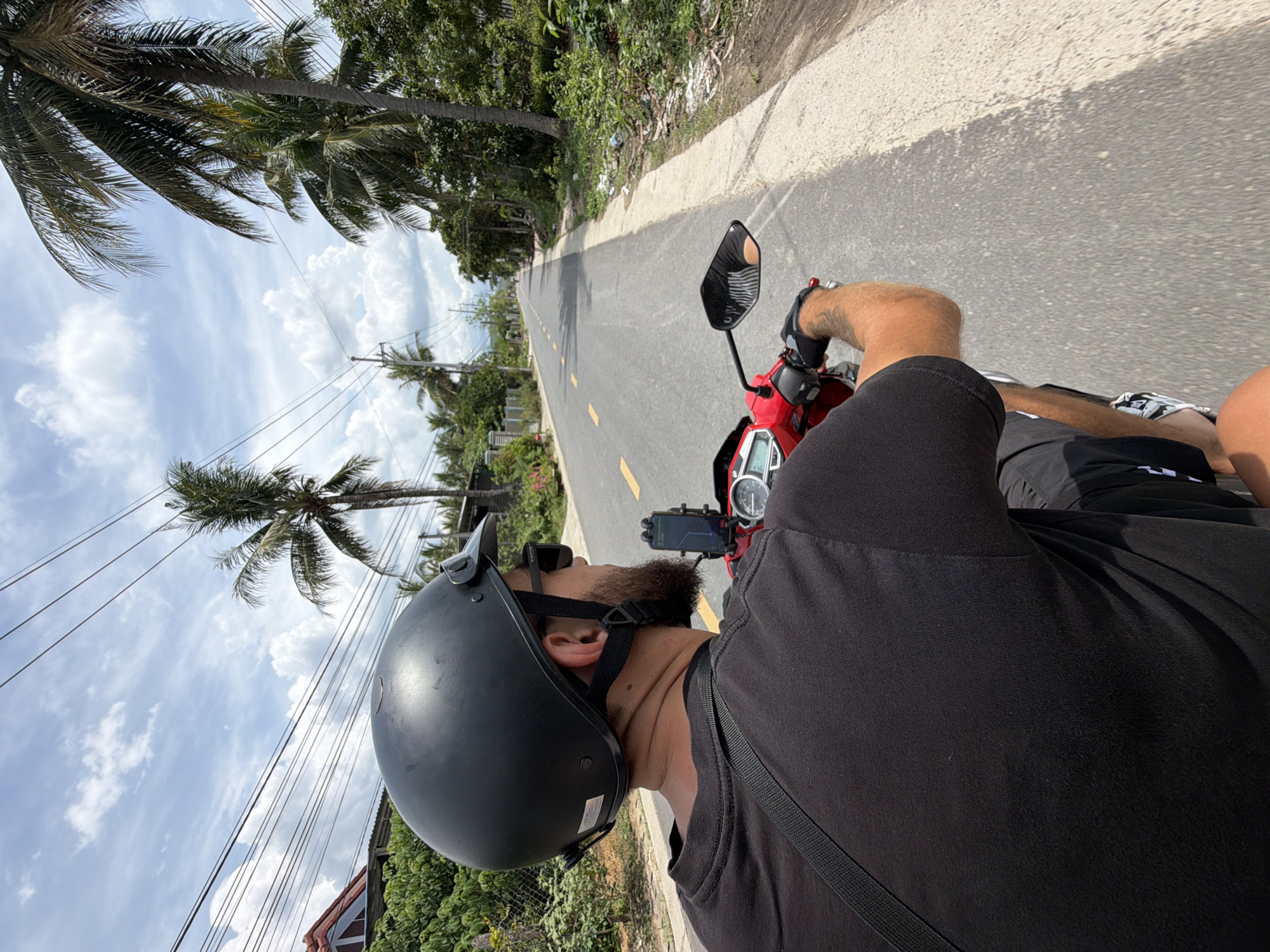 In an outdoor, daylight scene, a man wearing a black t-shirt and shorts rides a red scooter on a paved road lined with palm trees and lush vegetation. He is wearing a black helmet and sunglasses, and a black strap is visible across his chest. The road has yellow markings and appears to be in a rural or suburban area, with houses and power lines visible in the background. The sky is partly cloudy with scattered white clouds against a blue backdrop. The man is looking forward, and his hands are on the handlebars of the scooter. A phone is mounted on the handlebars. The location is identified as Diên Thọ, Vietnam.