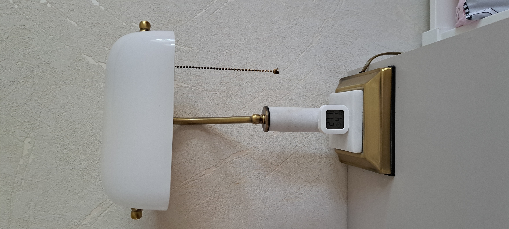 The image shows a close-up view of a wall-mounted light fixture. The fixture has a white, opaque shade and a brass arm extending from a brass base. A beaded pull chain hangs from the shade. Attached to the brass base is a small, white digital device with a screen displaying the numbers "21 41". This device appears to be a thermometer or a clock. The wall behind the fixture has a textured, light-colored wallpaper. To the right of the fixture, a portion of a white shelf or furniture is visible, with a corner of a patterned object on it, possibly a pillow or fabric. The lighting suggests it is daytime, and the setting appears to be indoors, likely a room in a house. No people are visible in the image.