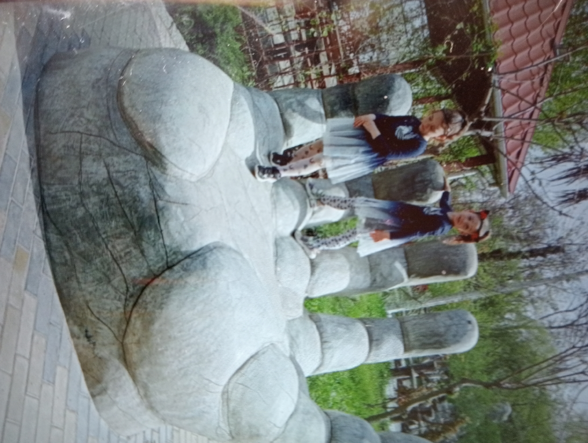 The image shows two young girls standing on a large, gray, sculpted hand in what appears to be an outdoor public space. The sculpture is textured to resemble skin and has distinct finger shapes. The girl on the left is wearing a dark long-sleeved shirt with a design on it, a light blue skirt, and patterned tights. She is looking at the camera. The girl on the right is wearing a dark blue long-sleeved shirt, a light blue skirt, and patterned tights. She is also looking towards the camera. Behind the girls and the sculpture, there are trees with green foliage and bare branches, and a red-tiled roof of a building is visible. The ground around the sculpture appears to be paved with light-colored bricks or stones. The lighting suggests it is daytime. The overall impression is of a playful moment captured in an artistic setting.