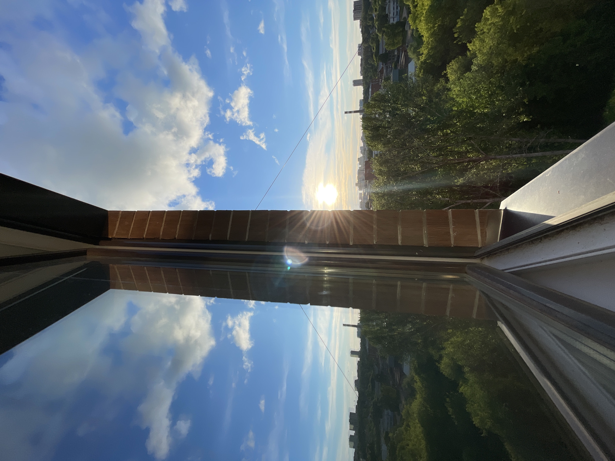 The image depicts a view from a window, likely in Izhevsk, Russia, showing a reflection of the sky with clouds and a cityscape in the distance. The window frame is visible, with brickwork along the upper part. The sun is visible, creating a lens flare effect. The scene shows a blend of urban and natural elements with buildings and green trees.