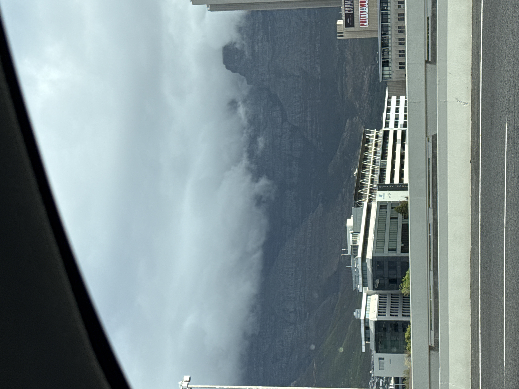 The image shows Table Mountain in Cape Town, South Africa, partially obscured by clouds. To the right are buildings including one advertising "PRETTY WOM". The view appears to be from inside a vehicle, with part of the interior visible on the left, and the highway visible to the right of the buildings.