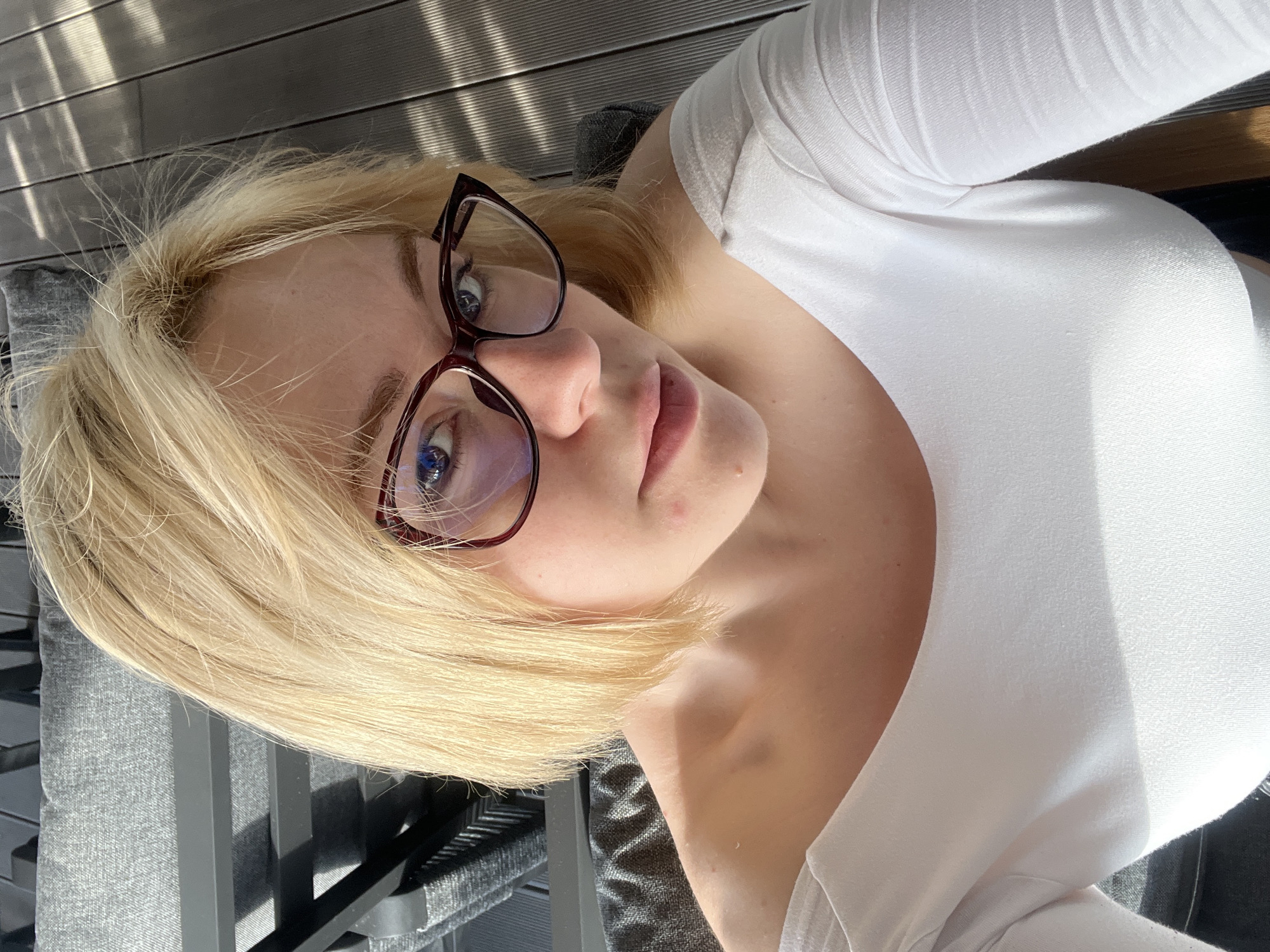 A young woman with blonde hair and blue eyes, wearing glasses and a white top, is seated outdoors. The location is Rafailovići, Montenegro, as suggested by the presence of outdoor seating structures similar to those found in coastal areas. The woman is positioned at an angle, looking directly at the camera. She has fair skin and is wearing light makeup. The glasses have a dark red or burgundy frame. The background features wooden decking or flooring with light reflecting off it, which is typical of seaside establishments. The outdoor seating appears to be made of gray metal or a similar material. The scene is well-lit, suggesting it is daytime. There are no other people visible.