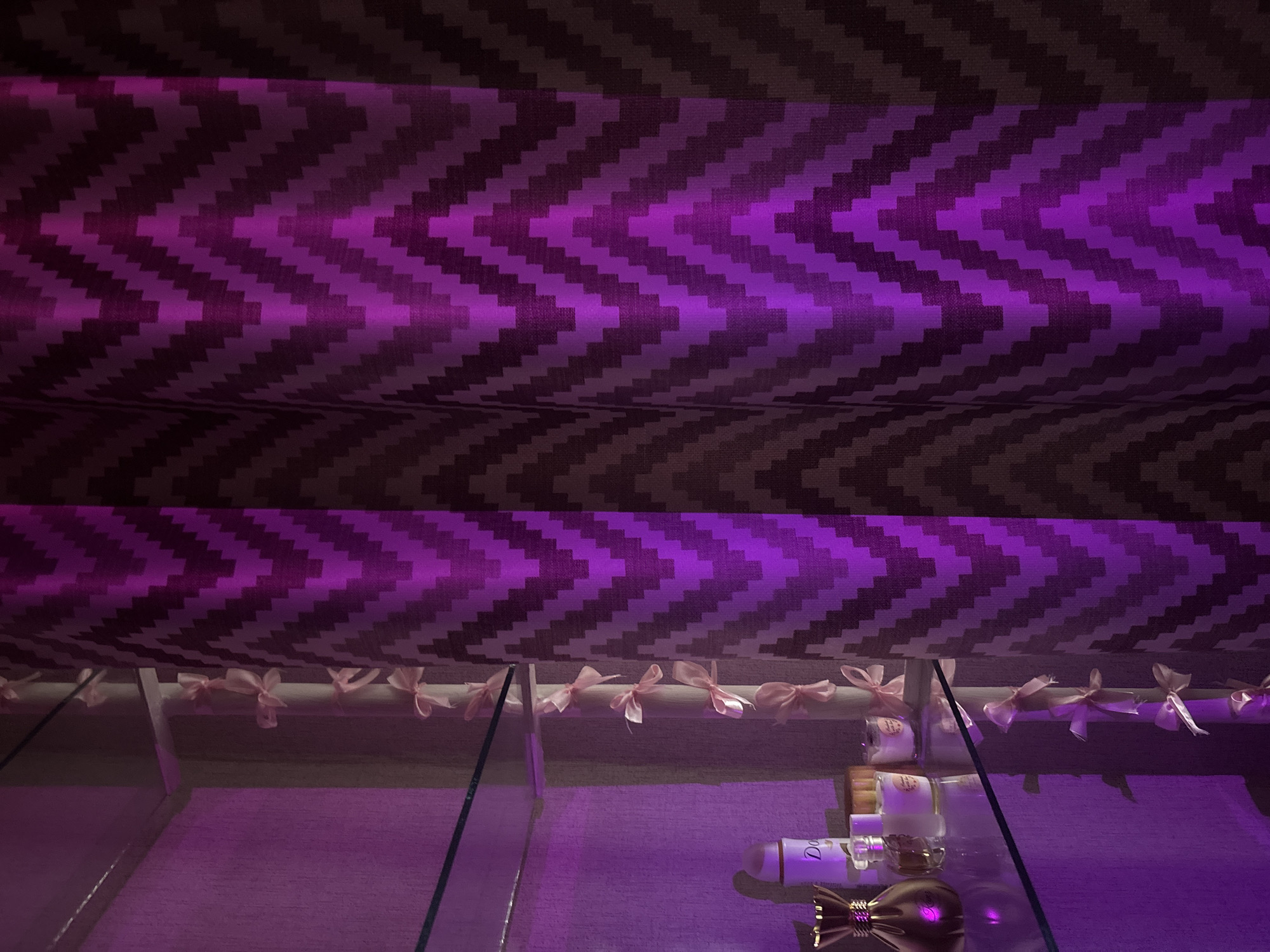 The image depicts a shelf in a domestic setting, illuminated by purple lighting. On the shelf, there are several perfume bottles and a spray can. To the right of the shelf is a curtain with a chevron pattern in shades of purple and brown. A string of small pink bows is visible hanging vertically along the side of the shelf. There are no people present in the image. The scene is indoors and appears to be a personal space, possibly a vanity or dressing area. The location in Inskoy, Russia, is not discernable from the visual information provided in the image.