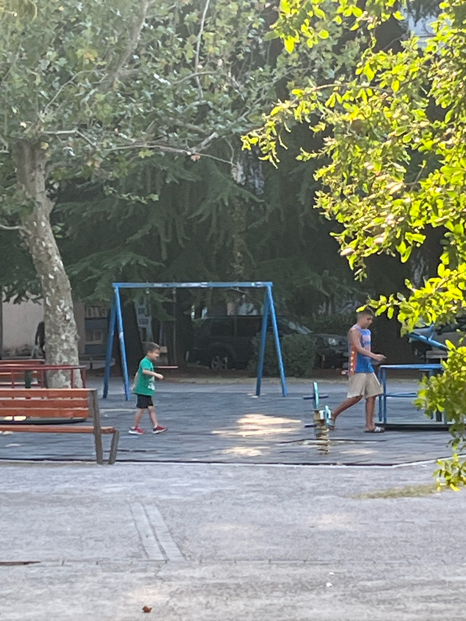 The image depicts an outdoor playground scene, likely in Podgorica, Montenegro, bathed in the warm light of what appears to be late afternoon or early morning. In the mid-ground, a young boy wearing a green t-shirt and black shorts is walking across the rubberized surface of the playground. To his right, an older boy or young man, dressed in a blue tank top with red and white accents and light-colored shorts, is also walking, his body angled away from the viewer. He is wearing sandals.
Dominating the background is a blue metal swing set with A-frame supports. Behind the swing set, trees with dense green foliage are visible, some branches extending into the foreground, casting dappled shadows. A portion of a park bench made of red wood and metal supports is present in the left foreground. The ground surface of the playground is a dark grey rubberized material, bordered by lighter grey paved areas. Shadows are cast across the ground, indicating a strong light source. The overall impression is one of a quiet, sunlit day at a public park.