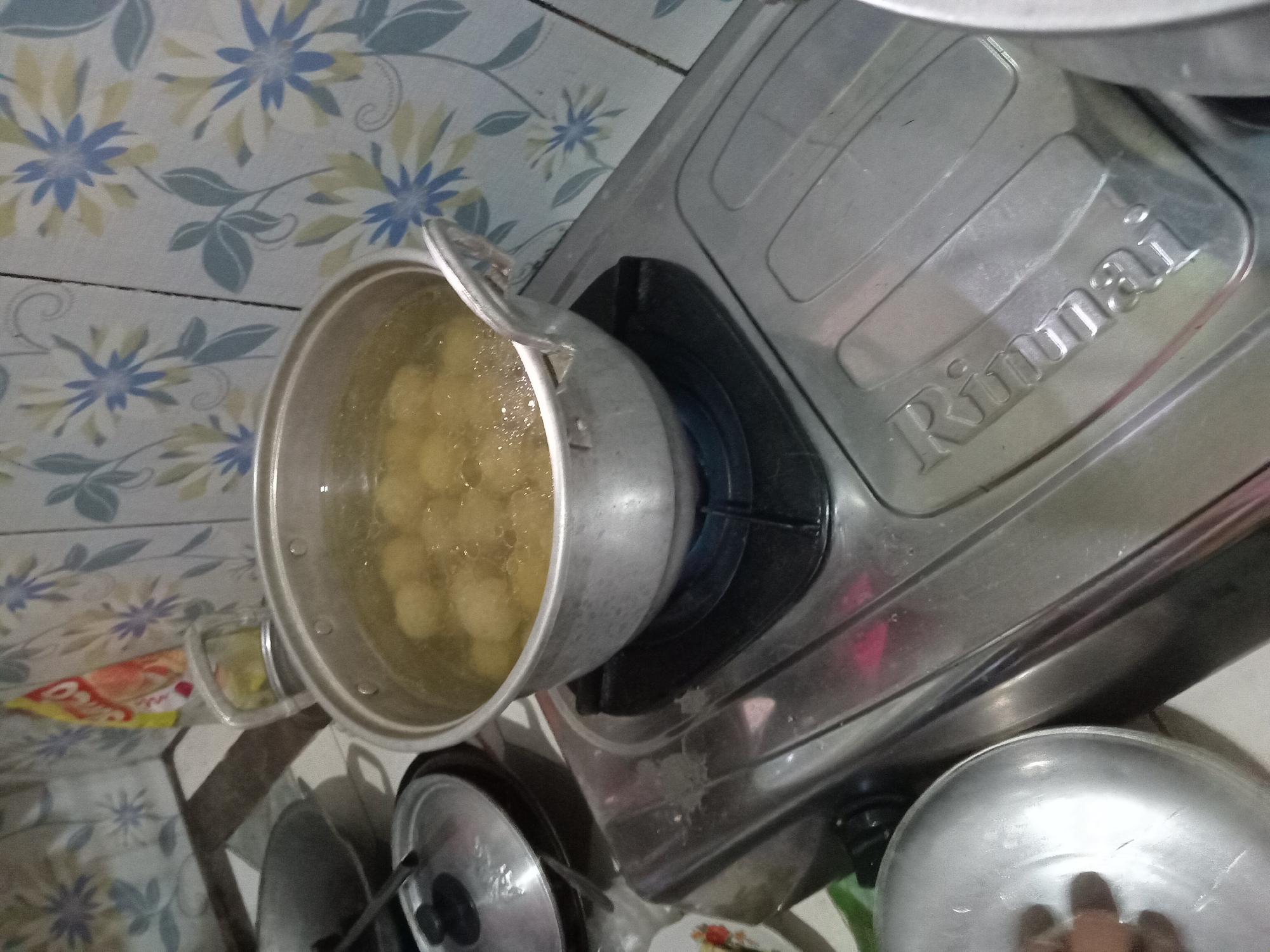 In Claket, Indonesia, a close-up shot captures a cooking scene. A metal pot filled with boiling water and dumplings sits on a gas stove. The stove, a Rinnai model, is made of stainless steel and shows signs of use. To the left of the stove, a covered pot and a stainless steel bowl are visible. The background features a wall tiled with a floral pattern in shades of blue, green, and white. The lighting is artificial and appears to be from overhead.