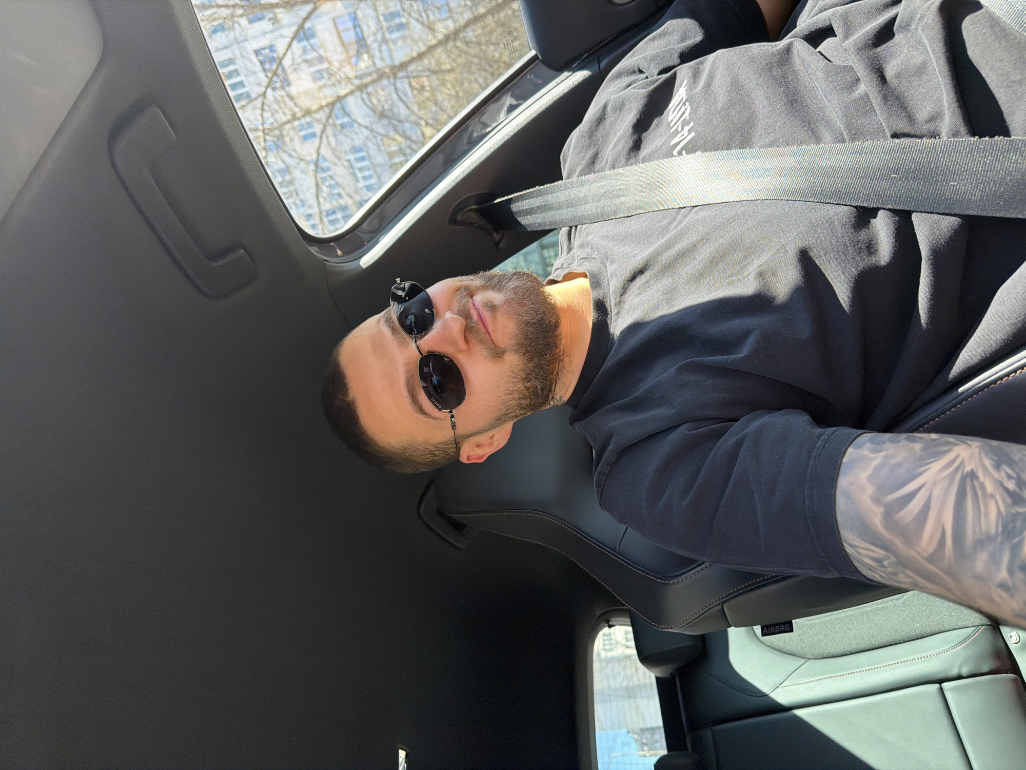 A man with a beard and sunglasses is sitting in the passenger seat of a car. He is wearing a black t-shirt and a seatbelt. The car has a sunroof, through which one can see a building and trees. The man has tattoos on his left arm. The location is Sofia, Bulgaria, indicated by the Cyrillic text on the t-shirt.