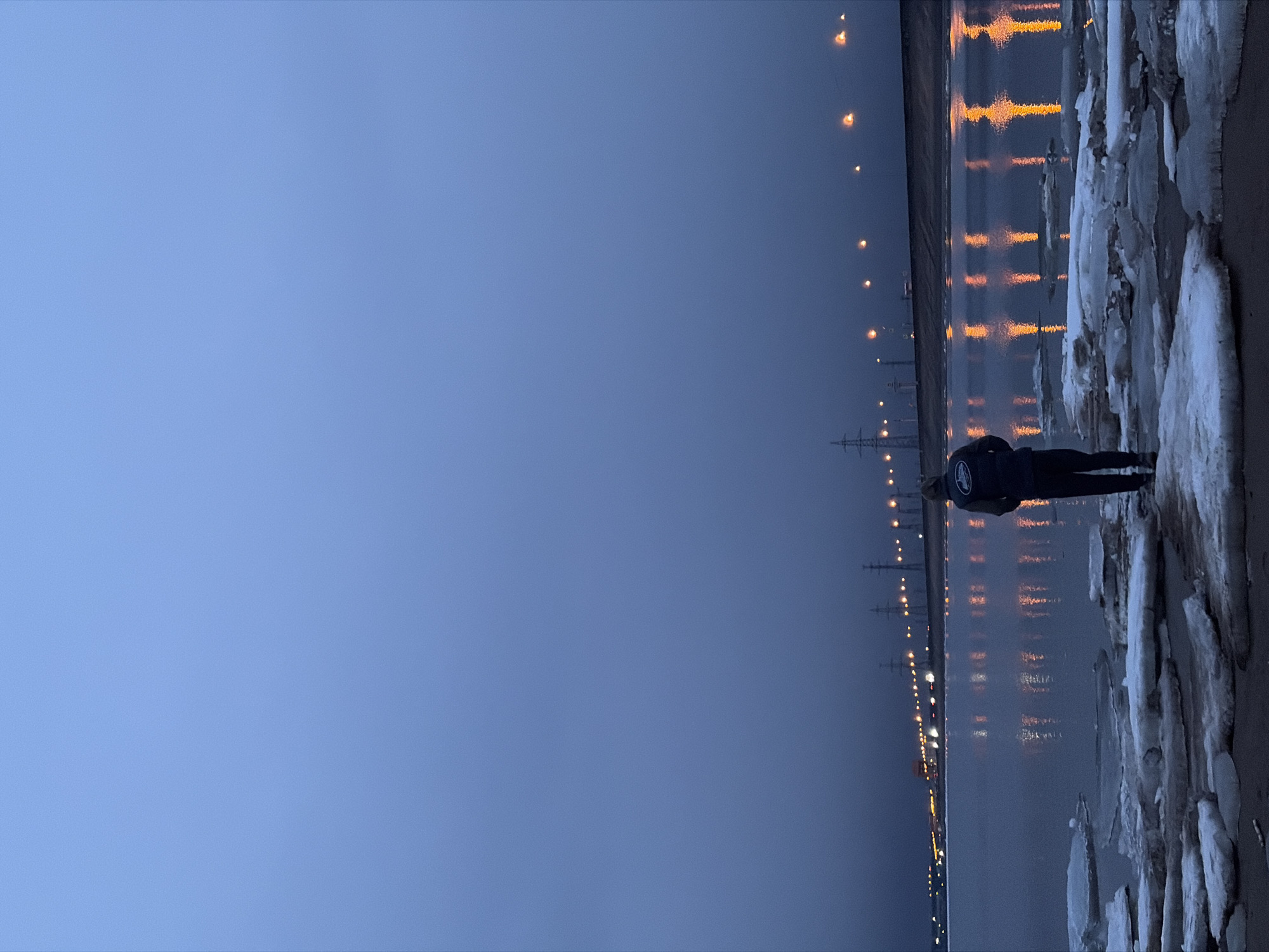 This image depicts a tranquil twilight scene by a large body of water in Noviy, Russia, likely during a cold period. The photograph is taken from a perspective looking across the water, with the horizon slightly tilted.
A single person, viewed from behind and silhouetted in dark clothing (possibly a jacket or hoodie with a circular logo on the back, though text is illegible), stands on the right edge of the frame. They are positioned on a bank covered with numerous irregular pieces of white and icy slush, indicating freezing temperatures.
The expansive body of water, which occupies the middle ground, reflects a series of bright, elongated streaks of golden-orange light. These reflections originate from a distant line of artificial light sources, possibly streetlights, situated along the far bank or shoreline to the left. Several power line towers or utility poles are faintly discernible against the dark horizon in the distance.
The sky, which dominates the upper and left portions of the image, is a deep, uniform blue-gray, characteristic of dusk or dawn. The overall lighting and visible ice suggest a cold climate and the transitional period between day and night when artificial illumination becomes prominent. The scene evokes a sense of quiet observation or solitude. No discernible text is clearly visible within the image.