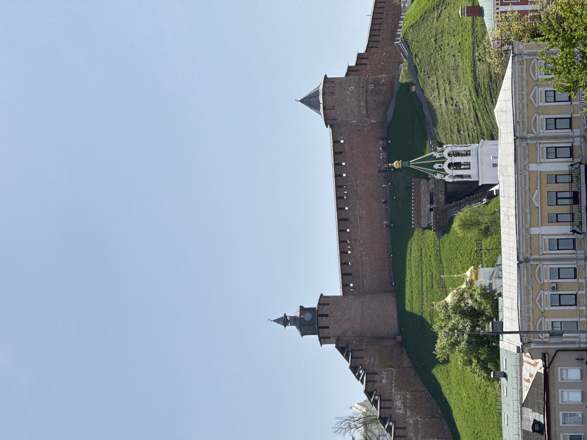 The image depicts a section of the Nizhny Novgorod Kremlin, a historic fortress located in Nizhny Novgorod, Russia. The view showcases the red brick wall with its towers rising along a green hillside. Adjacent to the wall, there are buildings with cream-colored facades and various architectural details. In the background, there is a clear, light blue sky, which occupies a large portion of the frame. The overall perspective suggests the photograph was taken from a low angle, looking upwards.