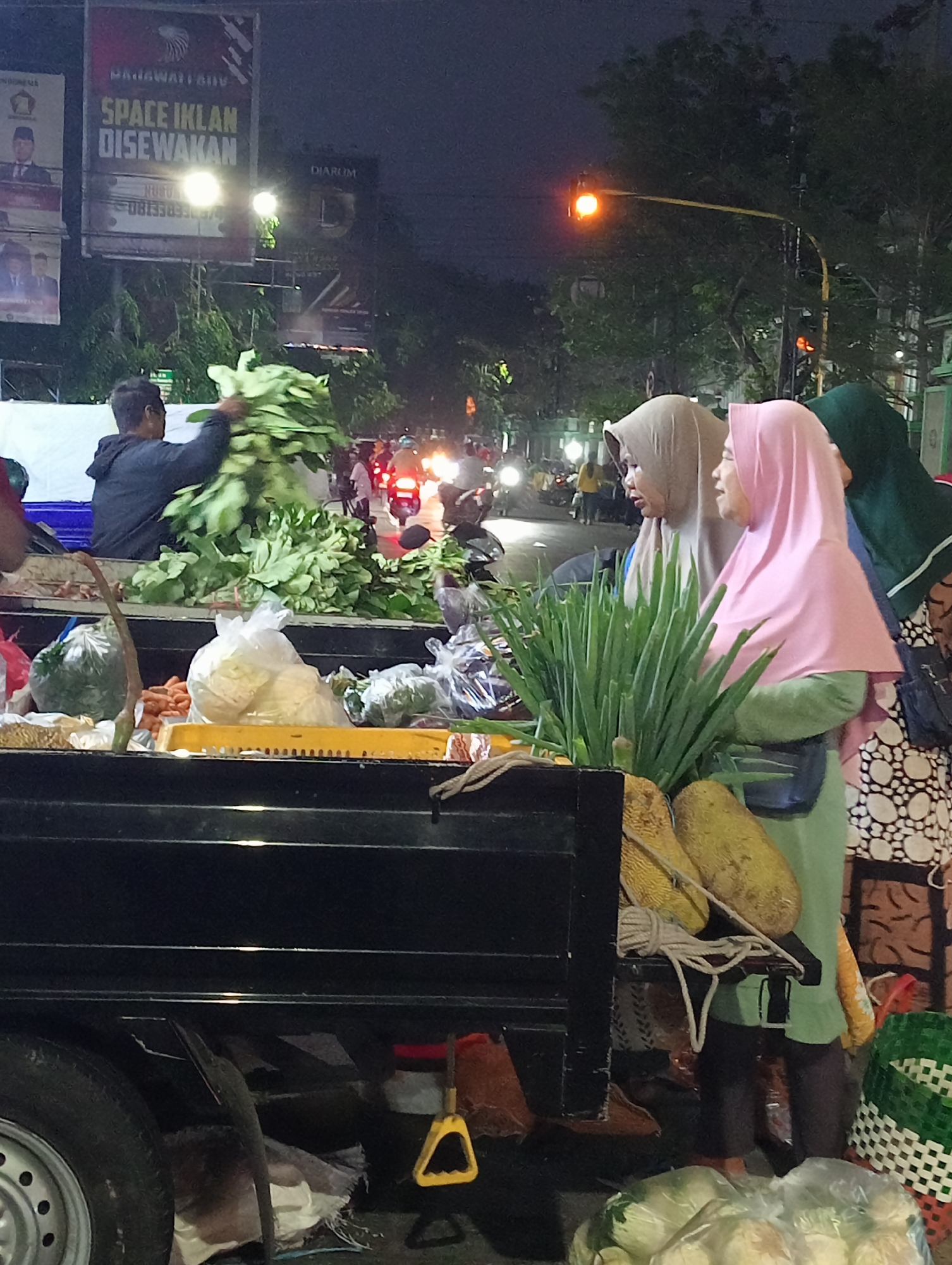 This image depicts a nighttime scene at a street market in Demak, Indonesia, likely near a busy intersection based on the traffic lights and visible signage. In the foreground, a black truck bed is laden with fresh produce, including large bunches of leafy green vegetables, plastic bags filled with cabbages, and carrots. Two women wearing hijabs are visible behind the produce; one in a light brown hijab and the other in a pink hijab, appearing to be interacting with each other or customers. To the left, a man in a dark hoodie is seen carrying a large bundle of green vegetables. In the background, streetlights illuminate the scene, and motorcycles and other vehicles are visible, suggesting ongoing traffic. A prominent billboard advertising "SPACE IKLAN DISEWAKAN" (Advertising Space for Rent) is lit up, with Indonesian text and logos. The overall atmosphere is that of an active night market.