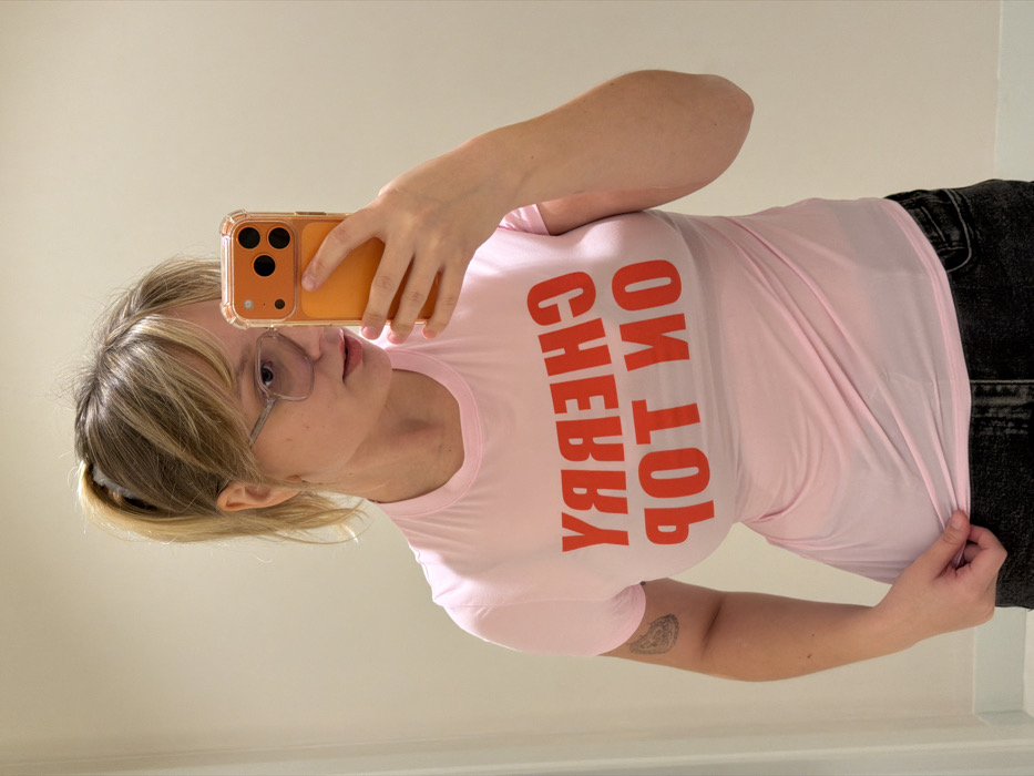 The image depicts a mirror selfie of a person, appearing to be a young adult female, standing indoors against a plain, light-beige wall. The specific location within Tel-Aviv, Israel, cannot be determined from the visual information provided.
The individual has blonde hair styled in a high ponytail secured with a light scrunchie, with bangs framing the face. She is wearing clear-framed glasses. Her right hand holds an orange-colored smartphone with a clear protective case, capturing her reflection. The phone's rear camera array is visible. She wears a light pink, short-sleeved t-shirt with the mirrored red text "CHERRY ON TOP" printed across the chest. The fabric of the t-shirt appears slightly sheer. A tattoo with a dark, intricate design is visible on her upper left arm. Her left hand lightly grips the bottom hem of her t-shirt. She is wearing dark-colored bottoms, possibly jeans, which are partially visible below the shirt. The environment features even, bright lighting and a simple, light-colored baseboard or floor at the bottom edge of the frame.