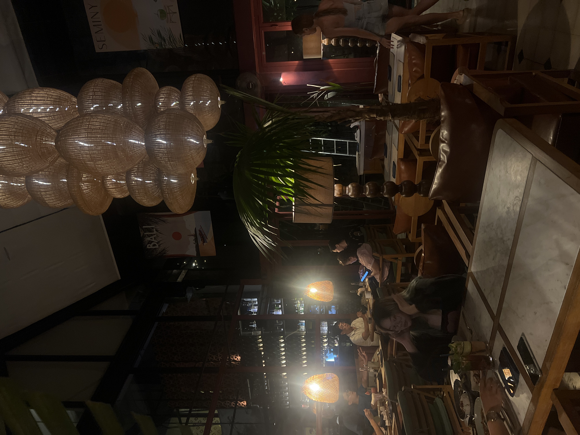 Captured from a slightly high angle, the photograph shows an evening scene inside a restaurant in North Kuta, Indonesia. Groups of patrons are seated at wooden tables and chairs, illuminated by warm, dim lighting from overhead woven pendant lamps. A palm tree is centrally placed within the restaurant’s interior. The architectural structure of the space is defined by dark beams, and decorative posters reading "SEMINYA" and "BALI" adorn the walls. Overhead, a person walks by, seen through an open doorway.