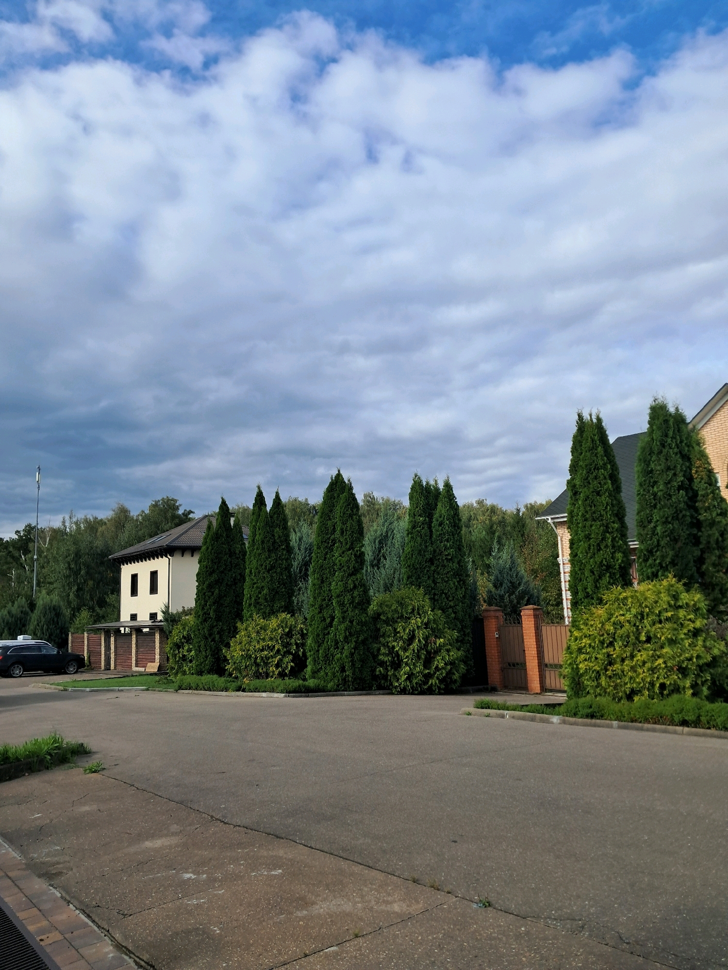 The image depicts a suburban street in Verkhneye Valuyevo, Russia, under a cloudy sky with patches of blue. A paved road occupies the foreground, with a concrete sidewalk along the left edge. To the right of the road, a neatly landscaped area features a row of tall, manicured evergreen trees and shrubs. Behind these, a brick gate and fence partially enclose a property. Further back, a light-colored house with dark windows is visible, with more trees and vegetation surrounding it. On the far left of the image, a dark-colored SUV is parked on the side of the road. The overall scene suggests a residential area with well-maintained properties and lush greenery.
