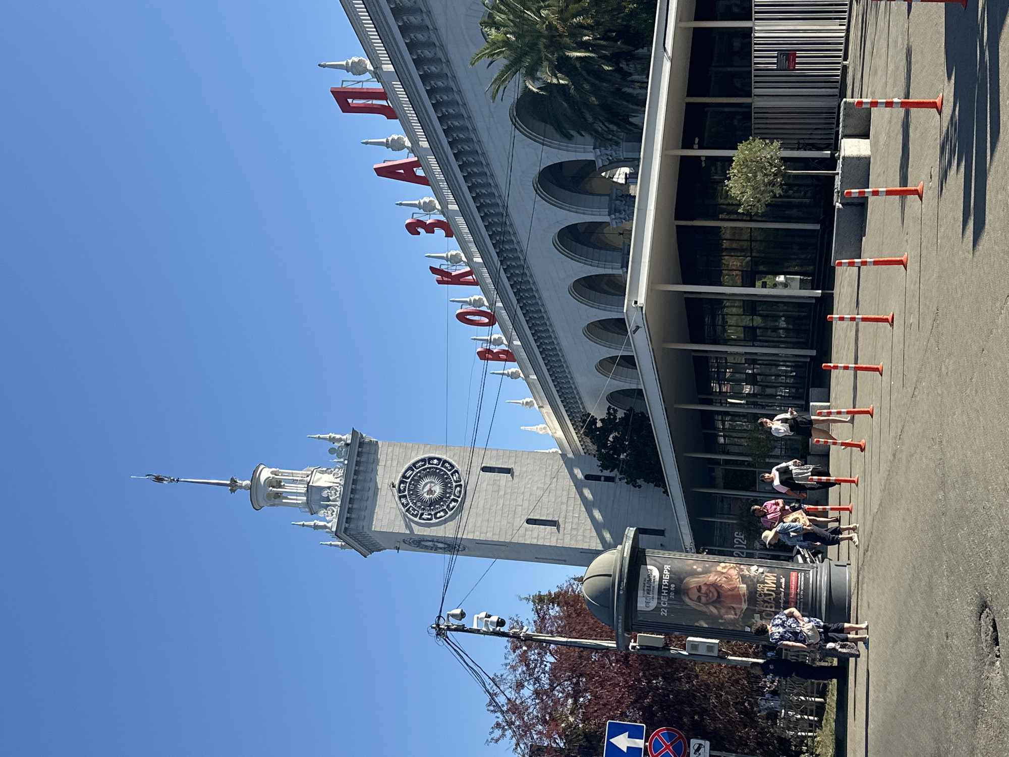 The image shows the train station in Sochi, Russia, on a clear, sunny day. The prominent features are the station building with its distinctive clock tower and the large red Cyrillic lettering "ВОКЗАЛ" (VOKZAL), meaning "train station". The architecture is grand, with arches and decorative elements. In the foreground, there is a street with a few people walking. A large advertising pillar stands to the left, displaying a poster. Orange and white traffic cones are arranged in a line on the asphalt. Trees, including a palm tree, are visible, suggesting a warm climate. The sky is a vibrant, cloudless blue.