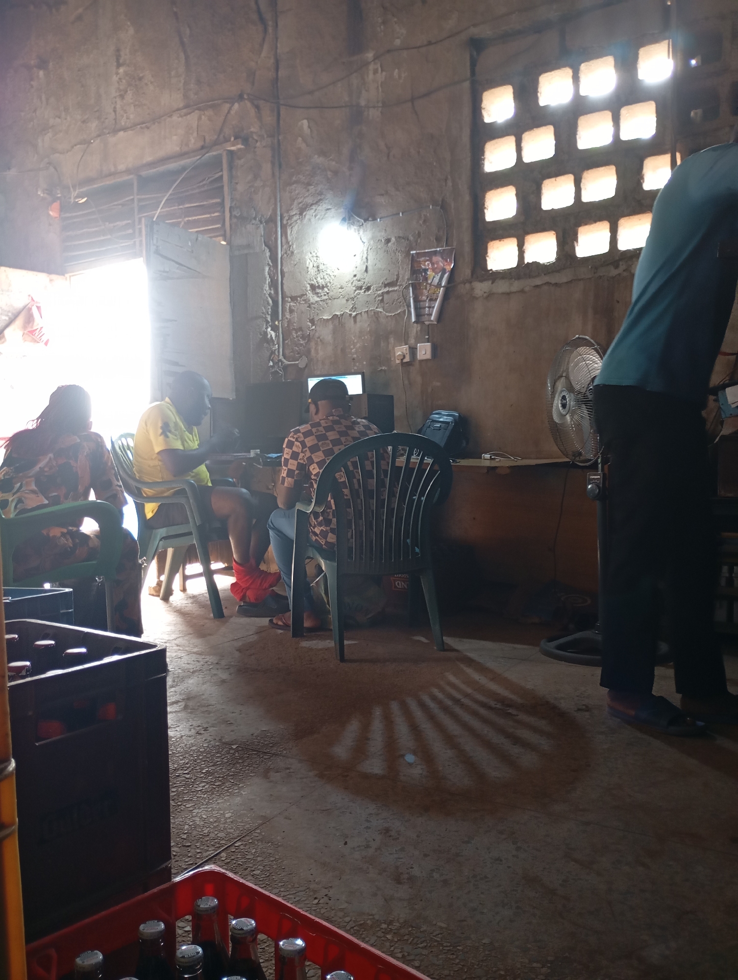 In Shendam, Nigeria, this interior scene depicts a rustic, open-air establishment with concrete walls and a corrugated metal roof. Sunlight streams through a large opening on the left, illuminating the space and casting long shadows from a chair on the floor. Several individuals are present. In the foreground, a person wearing a blue shirt and black pants stands, facing away from the camera. To their left, a stand fan is positioned. Further into the room, a man in a yellow shirt and dark shorts is seated at a table, gesturing with his hands. He is wearing red fabric wrapped around his legs. Across from him, another person in a checkered shirt and blue jeans is seated at the same table, looking towards the man in yellow. To the far left, a person with braided hair is seated, wearing a patterned top. On a table behind the seated individuals, a computer monitor and what appears to be a keyboard are visible. Various objects are scattered around, including plastic crates of bottles on the floor in the foreground, and a poster is affixed to the wall on the right. The overall impression is of a casual gathering or a place of business operating in a simple, open-air environment.