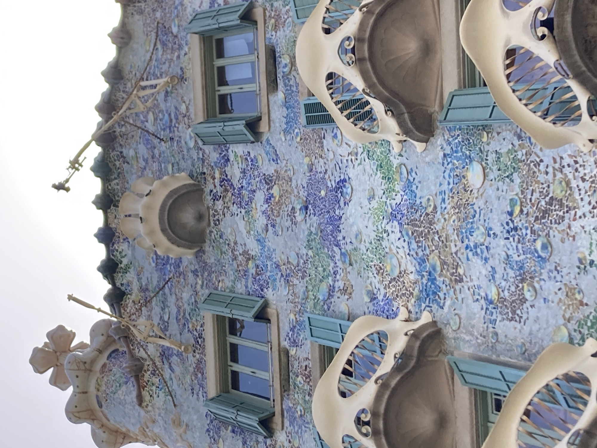 The image shows an upward-angled view of the facade of Casa Batlló, a building designed by Antoni Gaudí in Barcelona, Spain. The building's exterior is covered in a mosaic of colorful tiles, primarily in shades of blue, green, and white, interspersed with circular glass pieces that reflect the sky. The architecture features organic, undulating shapes, characteristic of Gaudí's modernist style.
Visible elements include several windows with light blue shutters. These shutters are partially open, revealing glimpses of the interior. There are also distinctive balconies with bone-like, white structures, creating an ethereal and somewhat skeletal appearance. One of these balconies has a spiral design. On the left side of the image, a unique, flower-shaped element protrudes from the facade, attached to a metallic arm or bracket.
The scene appears to be set during the daytime, with the bright sky suggesting good weather. The lighting indicates it is likely daytime, possibly late morning or afternoon, given the angle of the light. The vibrant colors and whimsical design of the building suggest a lively and artistic atmosphere. The image is taken from a low angle, emphasizing the scale and intricate detail of the architecture. There is no visible text in the image.