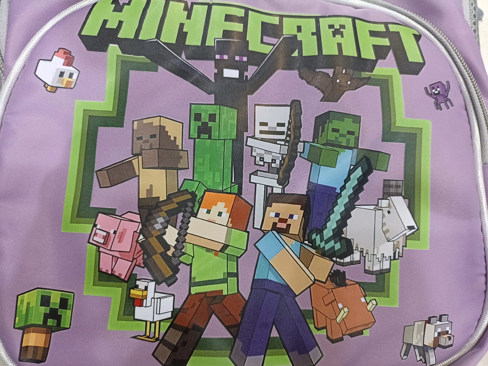 The image shows a close-up of a purple backpack with a Minecraft-themed graphic printed on it. The graphic features various Minecraft characters and creatures, including Steve, Alex, a Creeper, a Zombie, a Skeleton, an Enderman, a Pig, a Chicken, a Wolf, a Sheep, and a Bat. The characters are depicted in their iconic pixelated style, some holding weapons like swords and bows. The background of the graphic is a stylized green zig-zag pattern. The backpack itself has a white zipper and a textured gray strap. The image is well-lit and the graphic is clear and in focus. There is no discernible information in the image that would indicate its location in Mashhad, Iran, or any specific city.