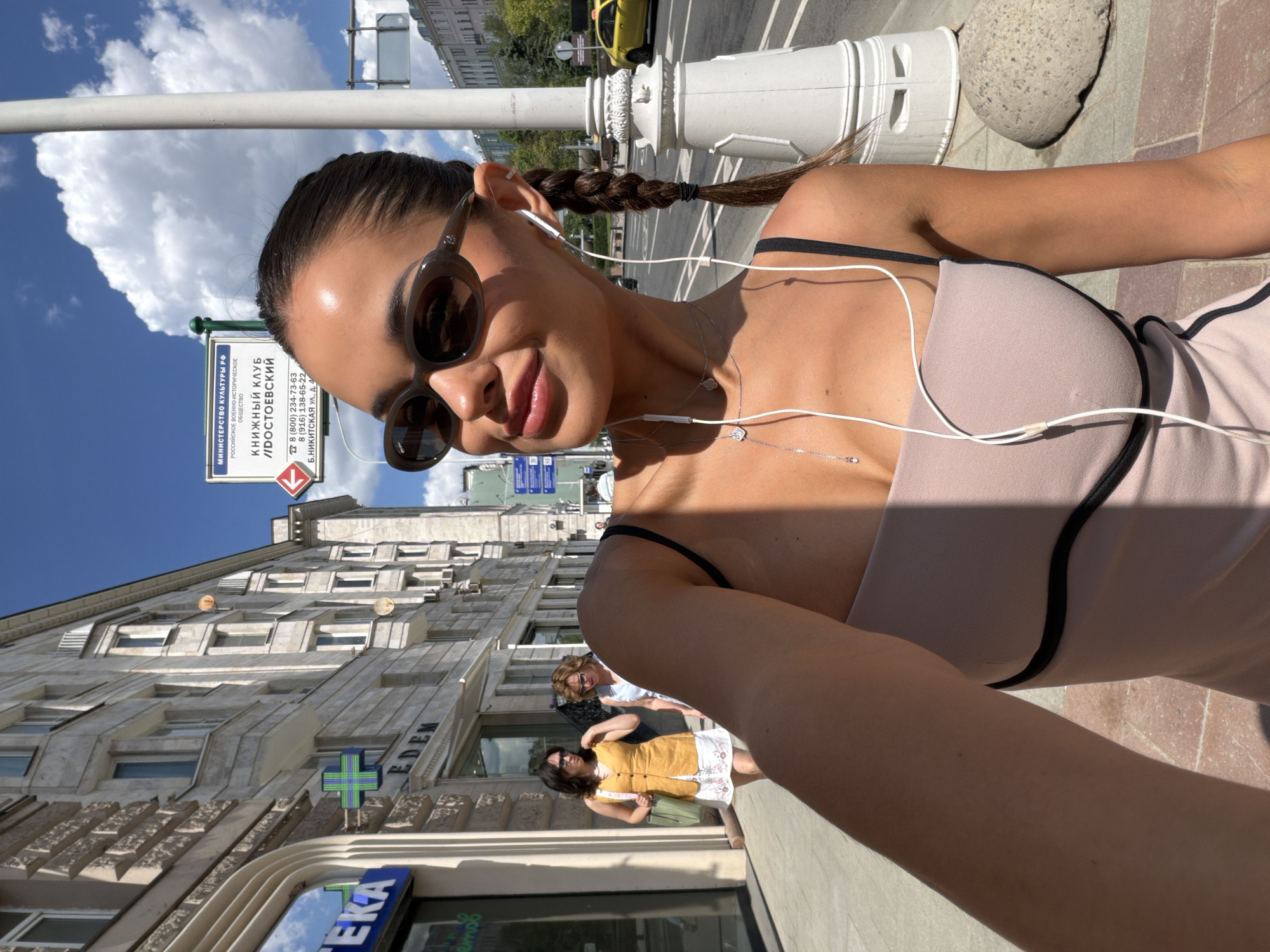 A young woman is featured in a close-up selfie taken on a sunny street, likely in Moscow, Russia. She has a tanned complexion, with dark hair neatly pulled back and styled in a braid that rests over her right shoulder. She wears dark, rounded sunglasses, white wired earphones, and a delicate silver necklace with small charms. Her light-colored top has black trim and visible black bra straps. She is smiling broadly towards the camera, conveying a pleasant mood.
The background reveals a vibrant urban street scene under a clear blue sky dotted with scattered white clouds. Tall, light-colored buildings with classic architectural details and numerous windows line the street. Several other pedestrians, including two women, are visible further down the sidewalk. A yellow taxi is partially seen in the background. A decorative white street pillar and a large cobblestone are also present on the sidewalk. Bright sunlight casts distinct shadows, indicating it is daytime.
A prominent blue street sign in the background displays significant Cyrillic text: "МИНИСТЕРСТВО КУЛЬТУРЫ РФ" (Ministry of Culture of the Russian Federation), "РОССИЙСКОЕ ВОЕННО-ИСТОРИЧЕСКОЕ ОБЩЕСТВО" (Russian Military-Historical Society), and "КНИЖНЫЙ КЛУБ //ДОСТОЕВСКИЙ" (Book Club //DOSTOEVSKY). The sign also lists phone numbers (8 (800) 234-73-63, 8 (916) 138-65-22) and an address: "Б.НИКИТСКАЯ УЛ., Д. 4" (B. Nikitskaya St., Building 4), confirming the location in Moscow. Additional visible text includes "EDEM" on a building facade and a partial reflection of "EKA" (likely from "APTEKA," meaning pharmacy) in a storefront window.