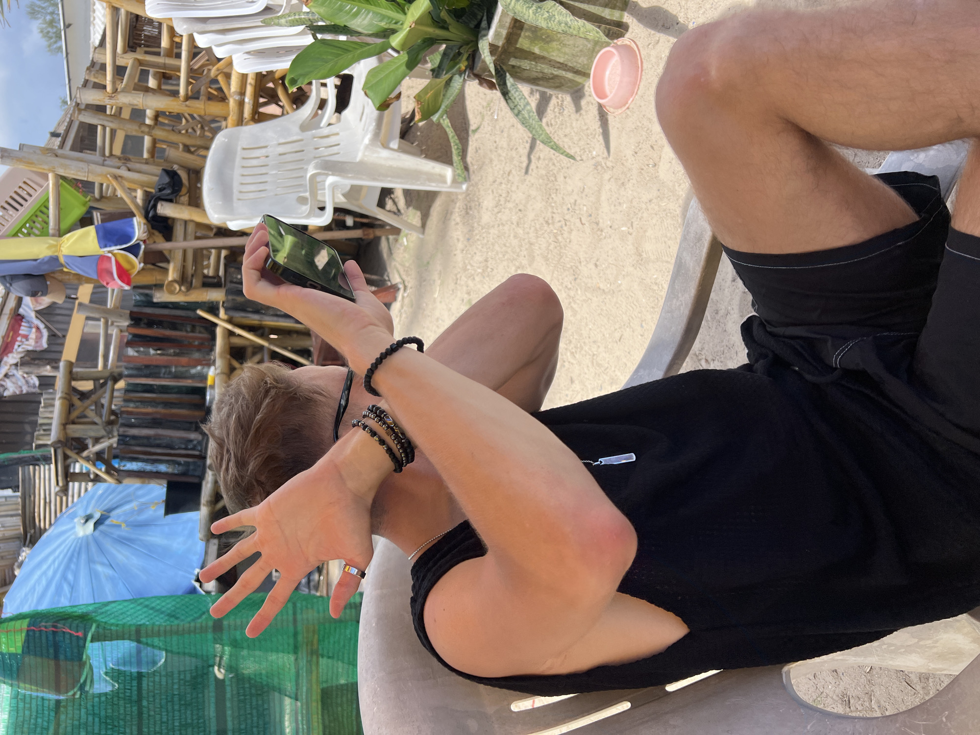 A young man, wearing a black sleeveless shirt and black shorts, is seated in a chair outdoors. He is holding a smartphone in his right hand, seemingly taking a picture or recording a video. His left hand is raised, with fingers spread, in front of his face, partially obscuring it. He is wearing a ring on his left ring finger and beaded bracelets on his left wrist. A silver pendant necklace is visible on his chest. The background shows a cluttered area with bamboo structures, white plastic chairs, a blue umbrella, and a potted plant. The ground is covered in sand.