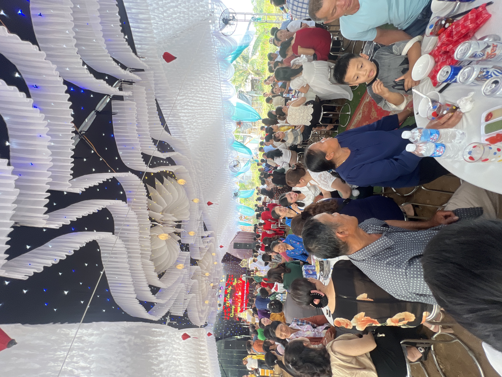 This media file captures a lively wedding reception held in a large, elaborately decorated tent or covered hall in Bình An, Vietnam. Numerous guests, comprising adults of various ages and some children, are seated at long tables, socializing and enjoying the festivities.
The most striking visual element is the overhead decoration, which features a dense arrangement of white, pleated fabric or paper structures, resembling clouds or stylized lanterns. These are illuminated by countless small, twinkling blue and white lights that mimic stars against a dark ceiling, creating a celestial and festive atmosphere. A few red, heart-shaped ornaments are also integrated into the decor. Further down the hall, large ceiling fans and some teal-colored translucent decorations are visible, suggesting an open-air or semi-outdoor setting.
Guests are seated at tables covered with white tablecloths. On the tables, bottled water, various brands of soft drink cans (such as Coca-Cola and 7-Up), and small plates with snacks or food are present. People are seen conversing, eating, and observing the event. A young boy in a grey shirt is visible near the foreground, looking towards the camera, while most attendees are oriented towards the far end of the hall.
At the distant end, a brightly lit stage or banner forms the focal point of the event. The banner, adorned with red and gold decorations, displays Vietnamese text. The legible text includes "Lễ Tân Hôn Ngọc" (Wedding Ceremony of Ngoc), "Quang," and "Ngày 8 tháng 3 năm 2023" (March 8, 2023), confirming the event as a wedding celebration for Ngoc and Quang on that date. The overall mood appears joyful and celebratory, with guests relaxed and engaged in the occasion. The lighting suggests the event might have transitioned from daytime into the evening.
