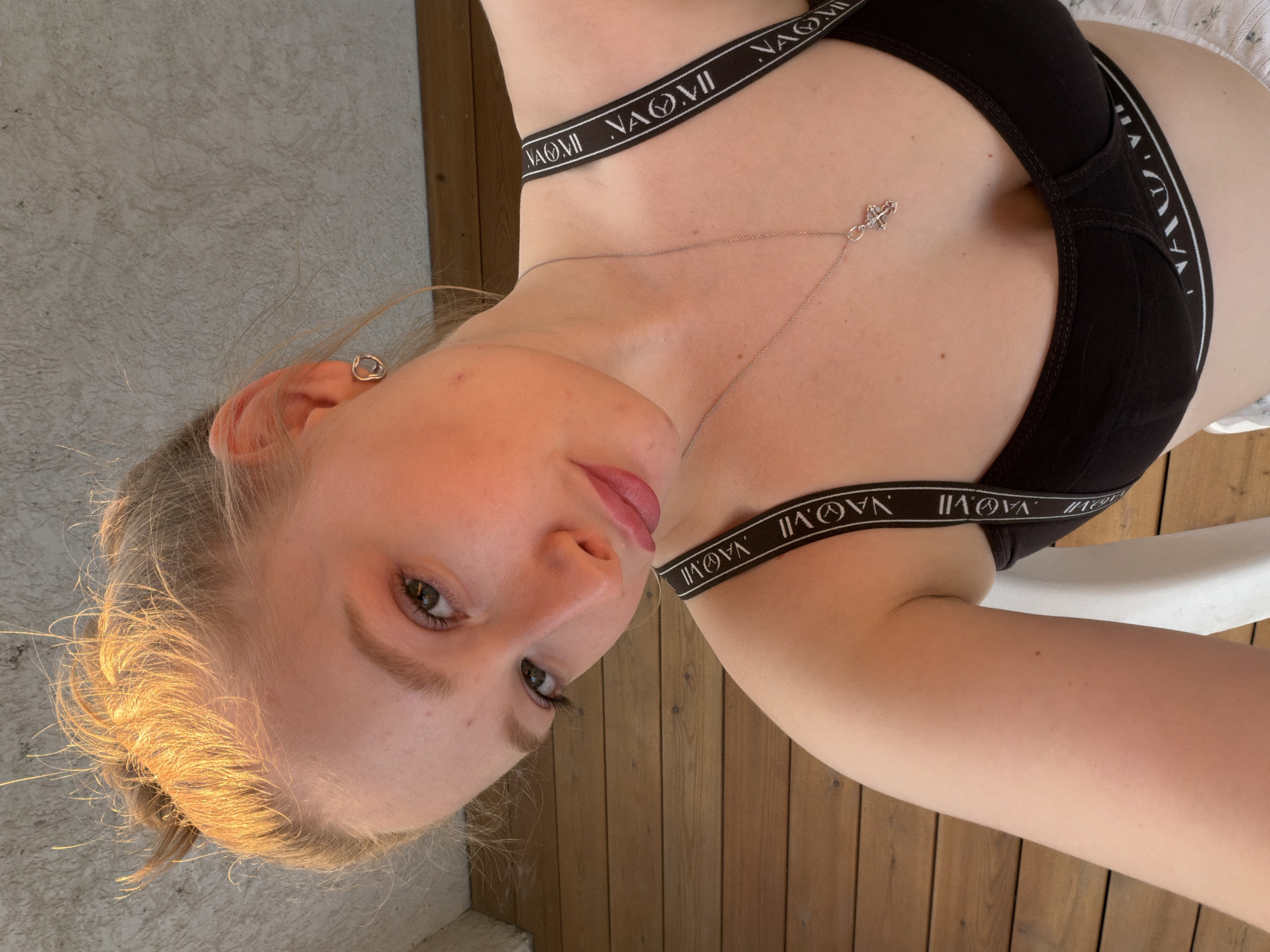 A young Caucasian woman with blonde hair pulled up in a bun is taking a selfie. She is wearing a black bra with a white logo that says "NAOMI VII" and a silver necklace with a cross pendant. Her makeup includes pink eyeshadow and lipstick. She has a heart-shaped earring in her left ear. The background shows a wooden deck and a textured wall. The lighting suggests it is either sunrise or sunset, casting a warm glow on her face. There is no discernible indication of her location within the city of Rossiyskiy, Russia.
