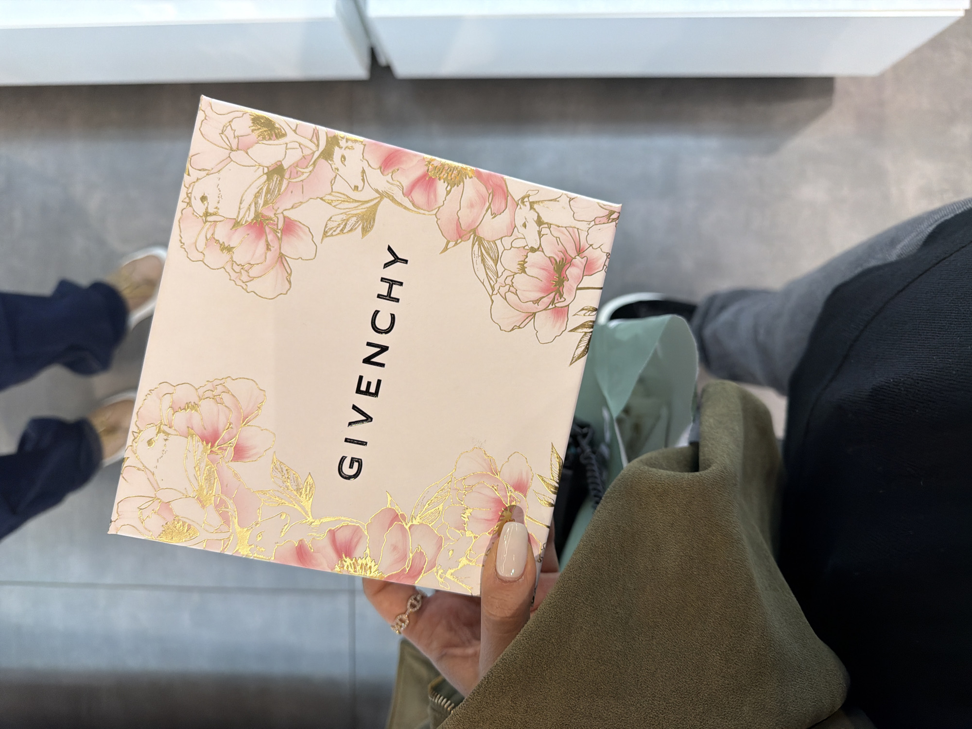 A person is holding a Givenchy branded box with a floral design. The box is rectangular and has a light pink background with pink and gold flowers framing the edges. The Givenchy logo is printed in black lettering down the middle of the box.
The person holding the box has manicured nails with a light neutral color. A gold ring is visible on one of the fingers. They are wearing an olive-green jacket, and parts of other people’s legs and feet are visible in the background. The surrounding environment appears to be a retail setting, possibly a department store, given the clean, tiled floor and the presence of other shoppers. There is no information to indicate that the location is Yerevan, Armenia.